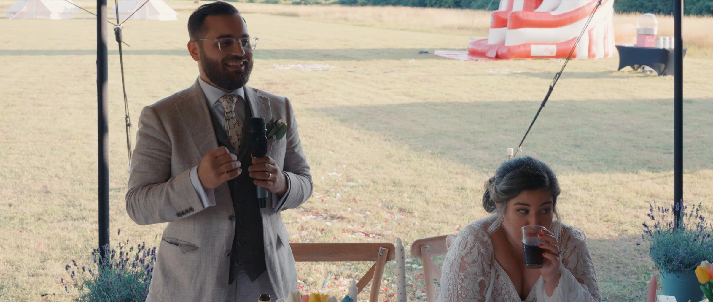 Man in a beige suit and glasses holding a microphone at an outdoor event, woman in a white lace dress sitting at a table drinking soda with a striped tent and field in the background.