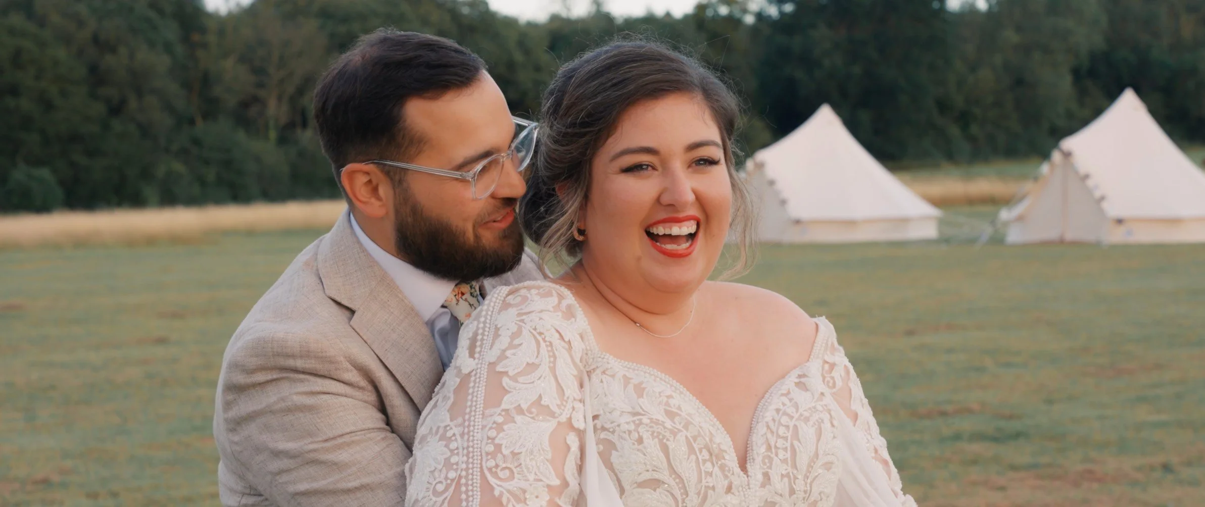 A couple dressed in wedding attire smiling and enjoying themselves outdoors near a body of water with tents in the background.