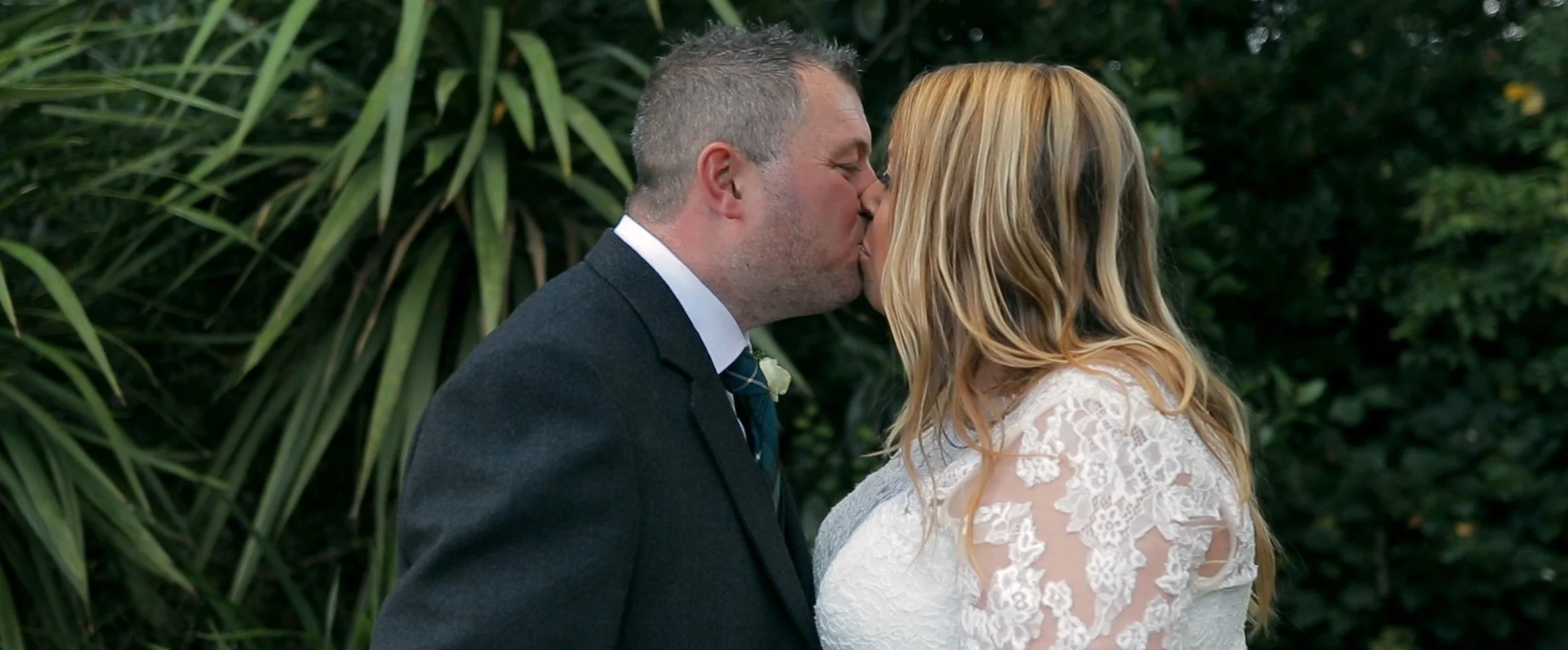 A couple in wedding attire sharing a kiss outdoors amid green foliage.