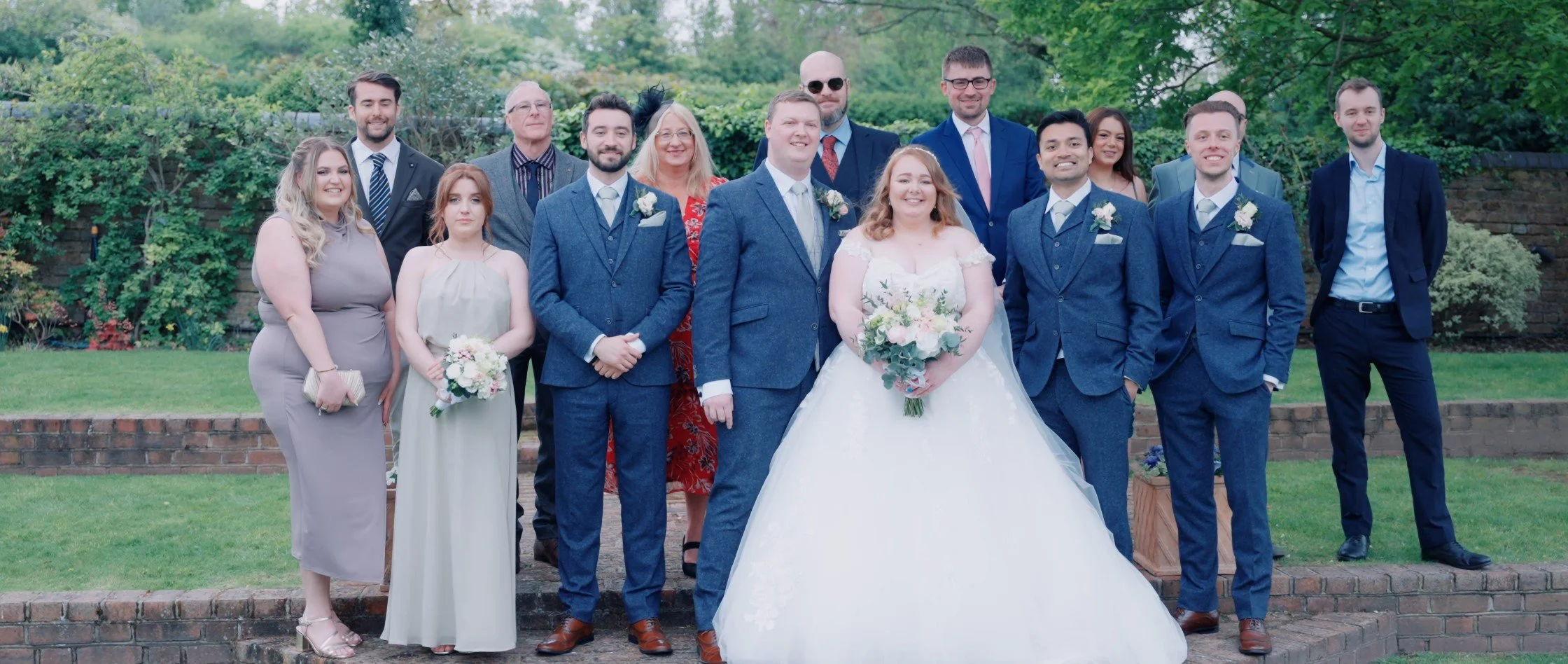 Group of people at a wedding, including the bride in a white gown holding a bouquet, and the groom in a blue suit, standing outdoors on brick stairs with greenery in the background.