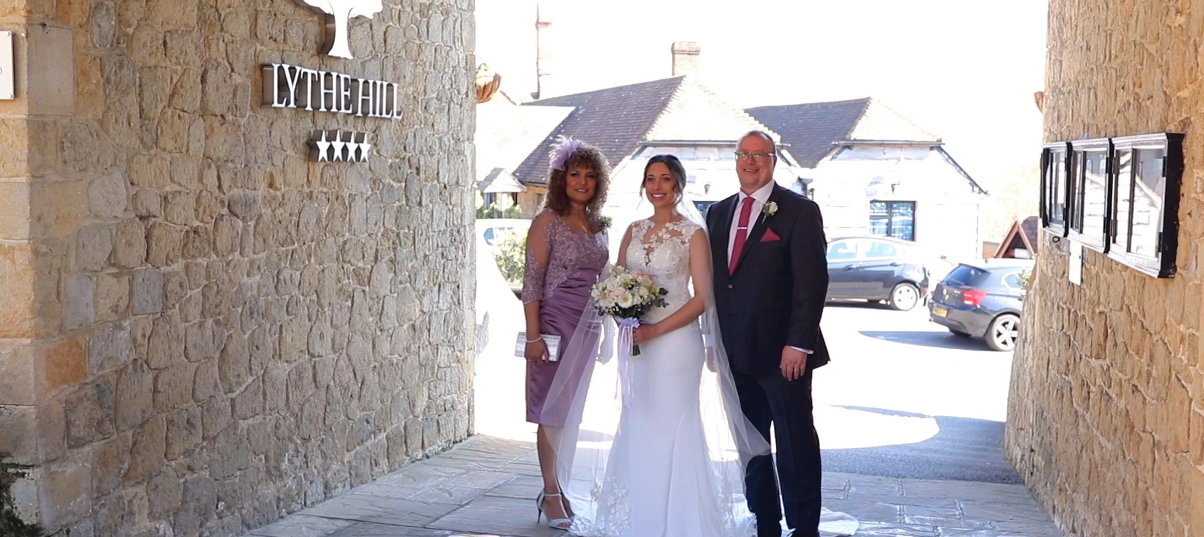 A bride in a white wedding dress holding a bouquet, flanked by a woman in lavender and a man in a black suit at the entrance of a stone building with a sign that reads "LYTHE HALL." The background shows parked cars and residential houses.