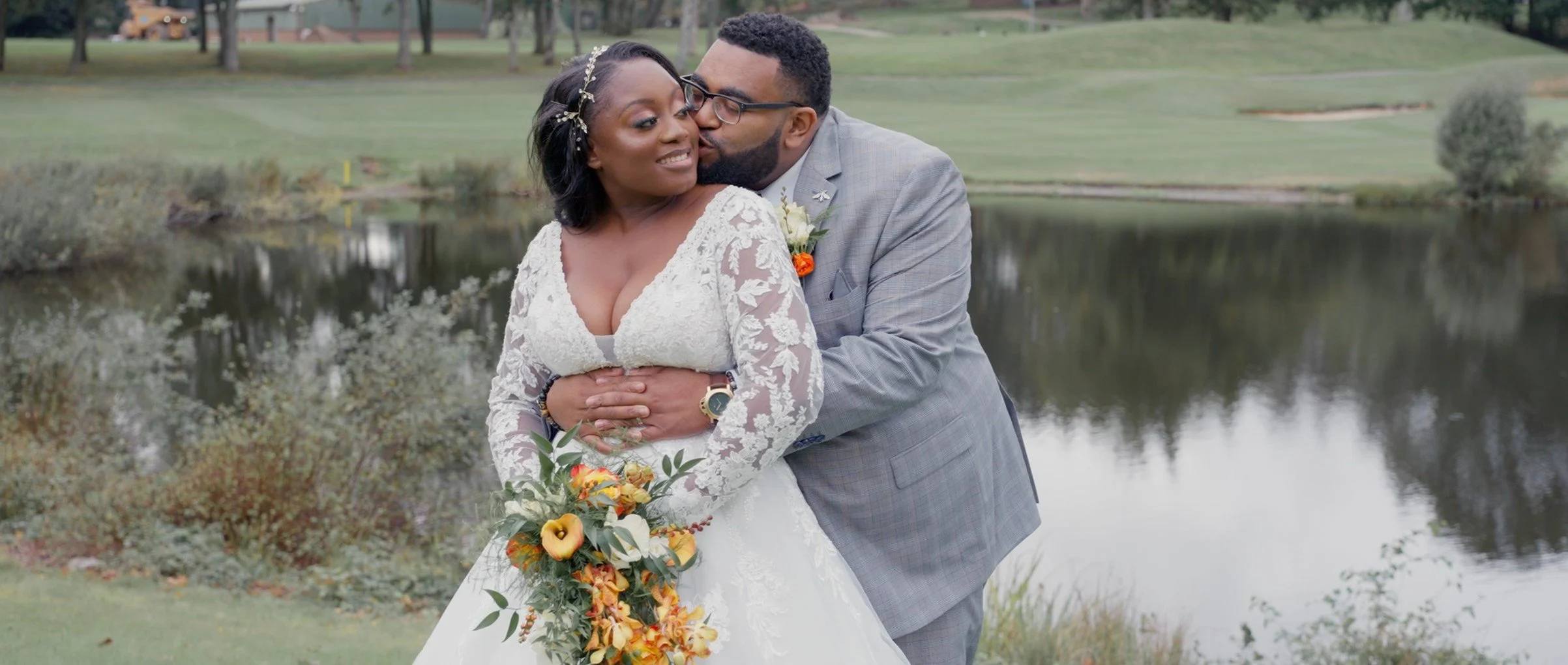 A bride and groom embracing by a lake in a park, with the groom kissing the bride on the cheek. The bride is holding a bouquet of orange and yellow flowers, wearing a white lace wedding dress. The groom is wearing a gray suit and glasses.