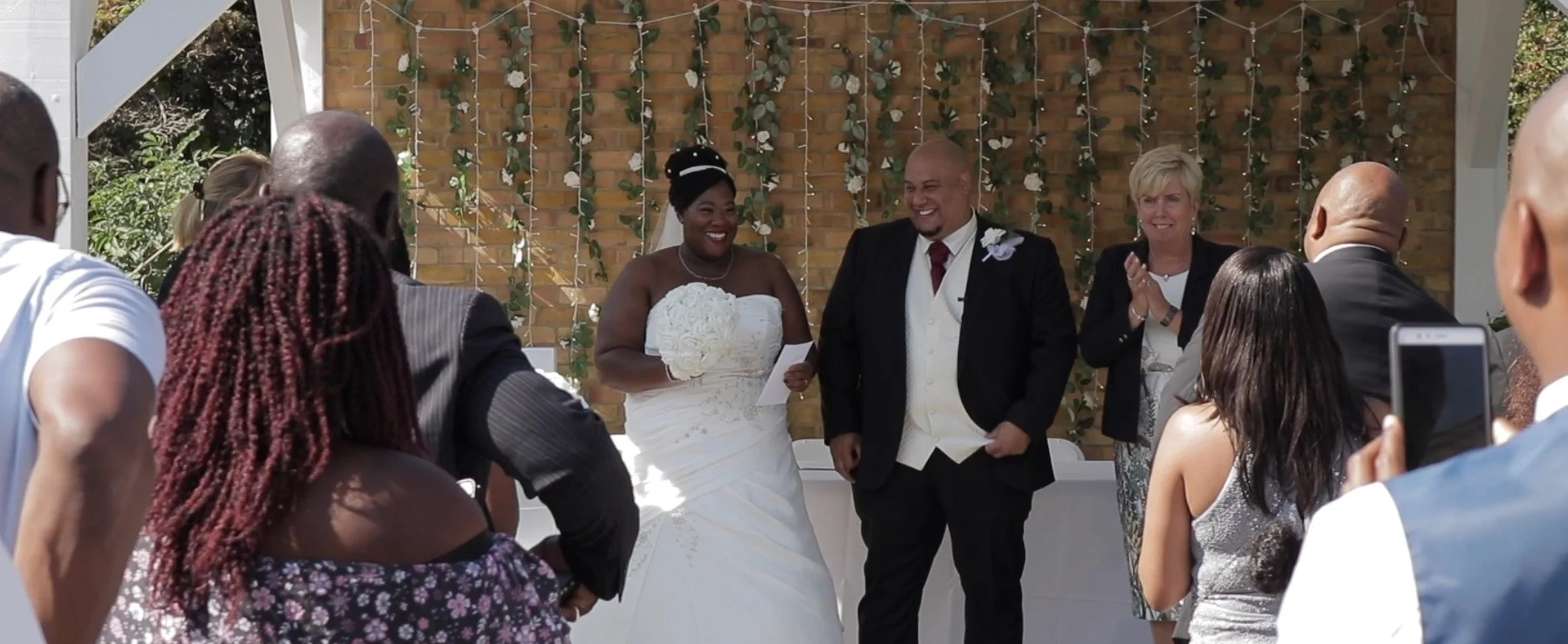 A bride and groom exchanging vows at their wedding ceremony, surrounded by friends and family, outdoors with a decorated brick wall and floral backdrop.