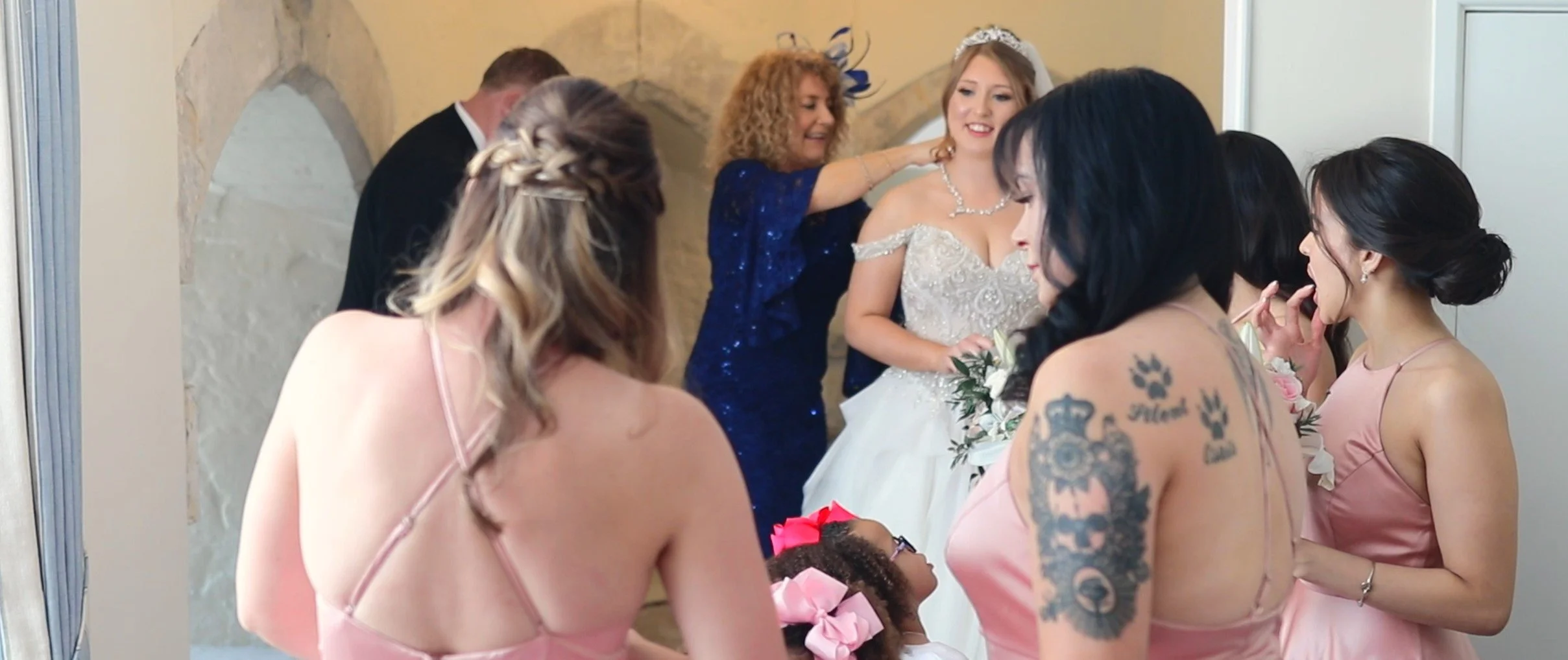 A bride in a wedding dress with jewelry is surrounded by bridesmaids. A woman in a blue dress adjusts the bride's earrings while others prepare for the wedding in a room with a beige wall and a decorative mirror.