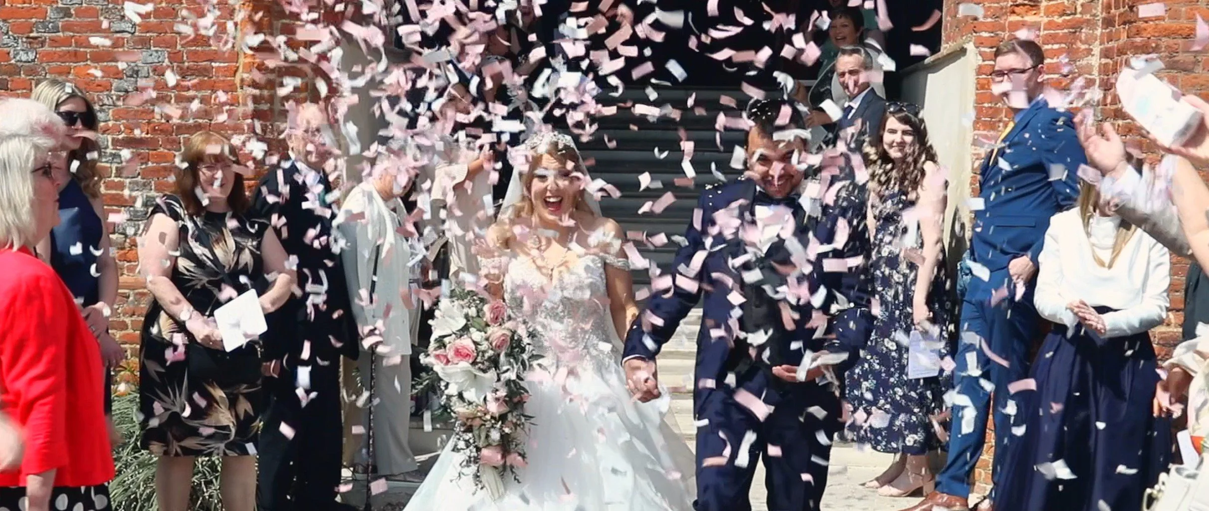 Bride and groom holding hands and smiling as pink and white confetti falls around them during their wedding celebration outside in front of a brick building, surrounded by friends and family.