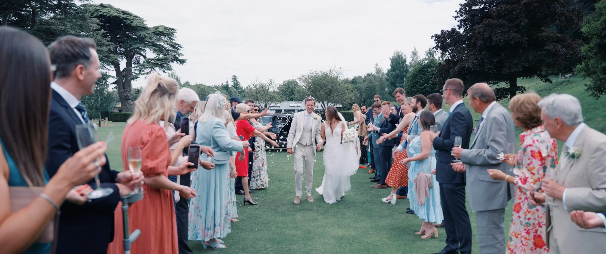 A wedding celebration outdoors with a bride and groom walking through a aisle created by guest lining on both sides, guests holding glasses of champagne and taking photos; trees and a grassy lawn in the background.