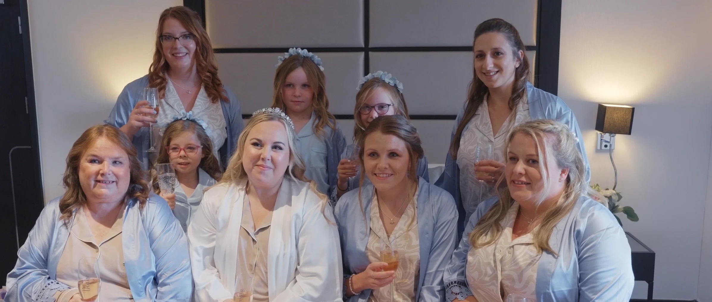 A group of women and girls wearing matching pajamas, some with floral headbands, gathered together for a celebration with champagne glasses in hand, smiling and posing for the photo in a room with modern decor and warm lighting.