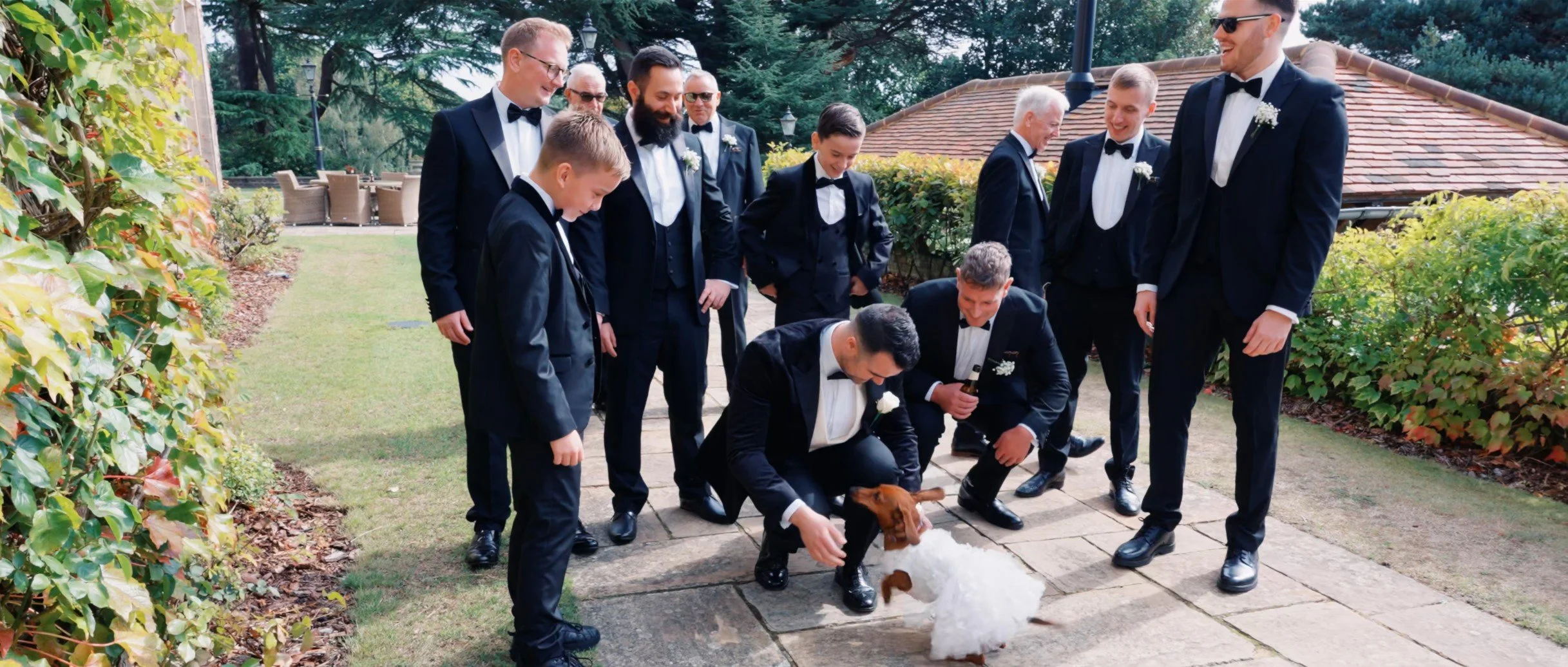 Group of men in black tuxedos with bow ties, some wearing sunglasses, gathered outdoors on a stone patio, as a man in tuxedo crouches to pet a small dog, surrounded by greenery and a building with a tiled roof in the background.