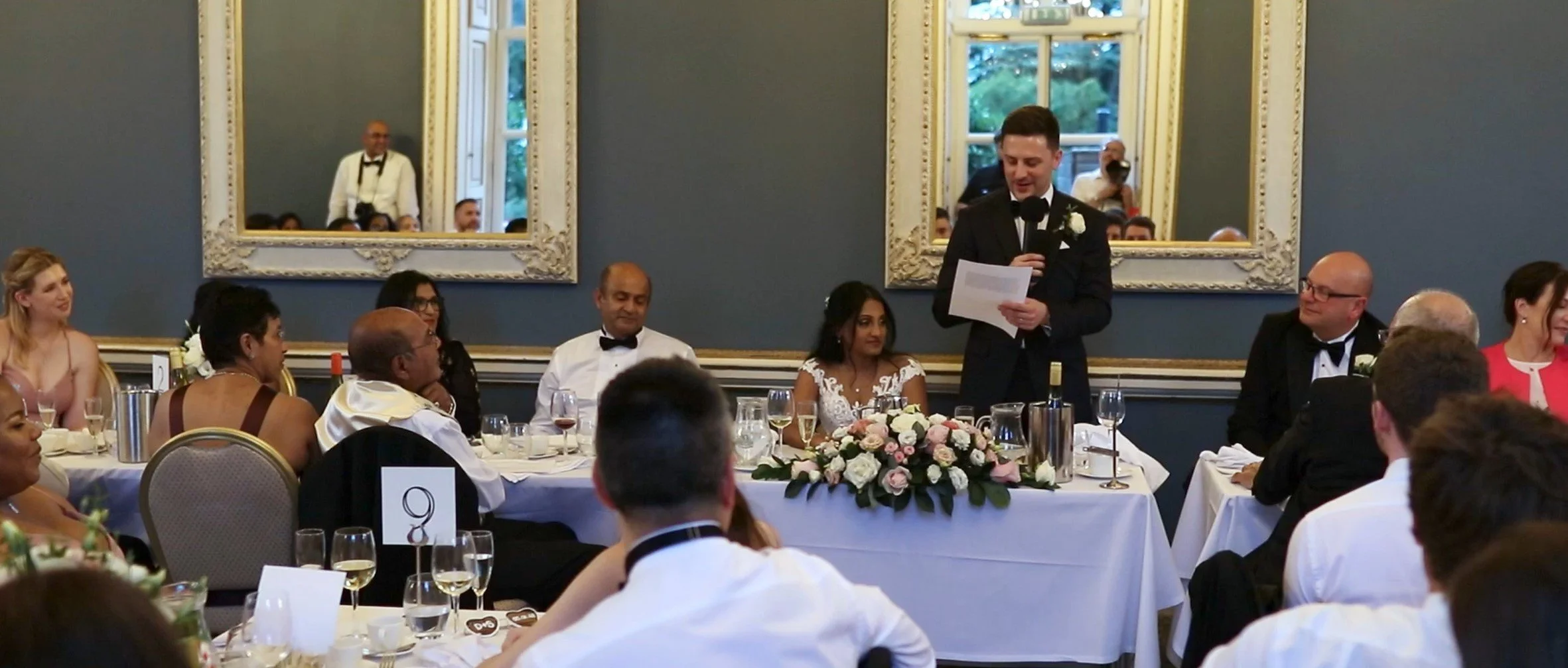 Wedding reception scene with guests seated at round tables, a man in a black suit giving a speech with a microphone, a floral centerpiece, and large ornate mirrors on a blue wall in the background.
