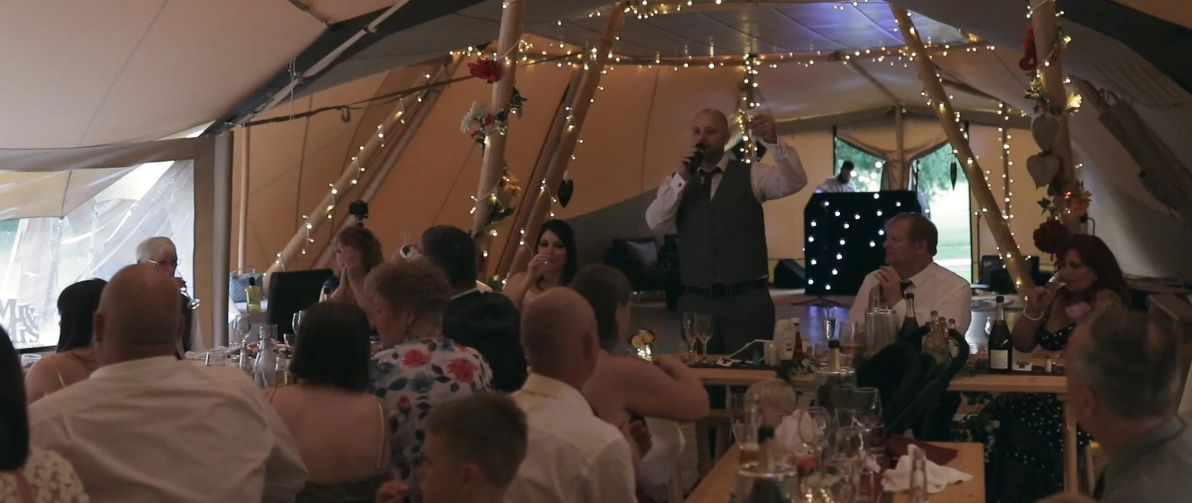 A man giving a speech at a wedding reception inside a decorated tent, surrounded by seated guests and string lights.