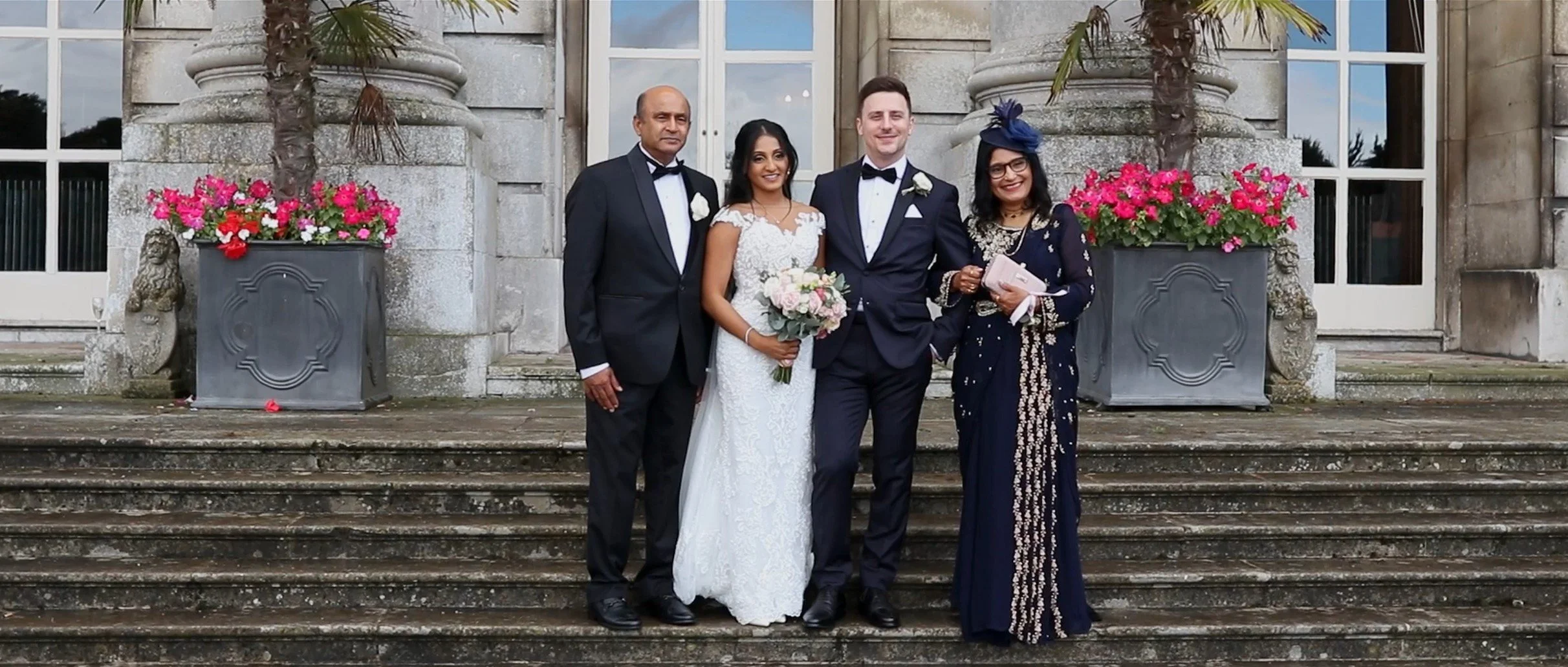 A group of five people, including a bride and groom, standing on steps in front of a stone building with large windows and pink flowers in large planters. The bride is holding a bouquet of pink and white roses, and everyone is dressed in formal attir