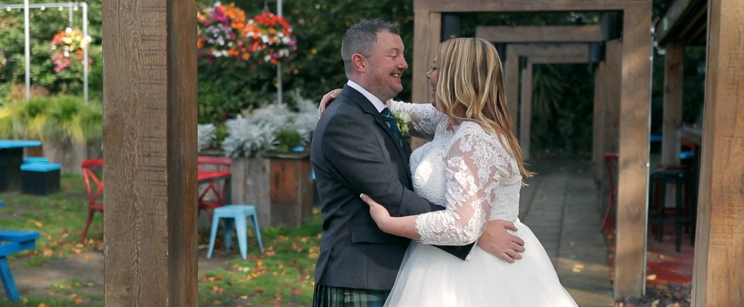 A bride and groom holding each other and smiling during their wedding celebration outdoors, with colorful flowers and decorative elements in the background.