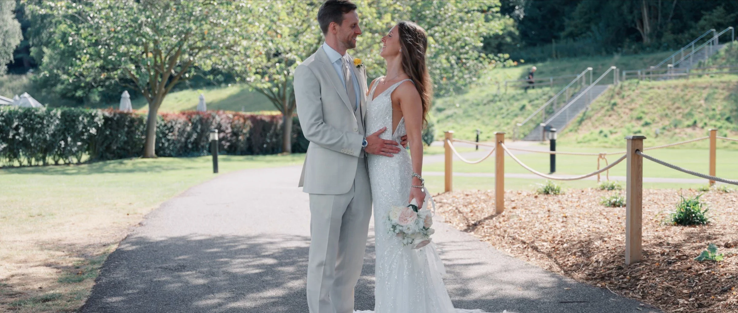 A bride and groom are standing close together outdoors during their wedding, smiling at each other. The bride is holding a bouquet of flowers and wearing a white gown, while the groom is dressed in a light-colored suit with a boutonniere. They are in