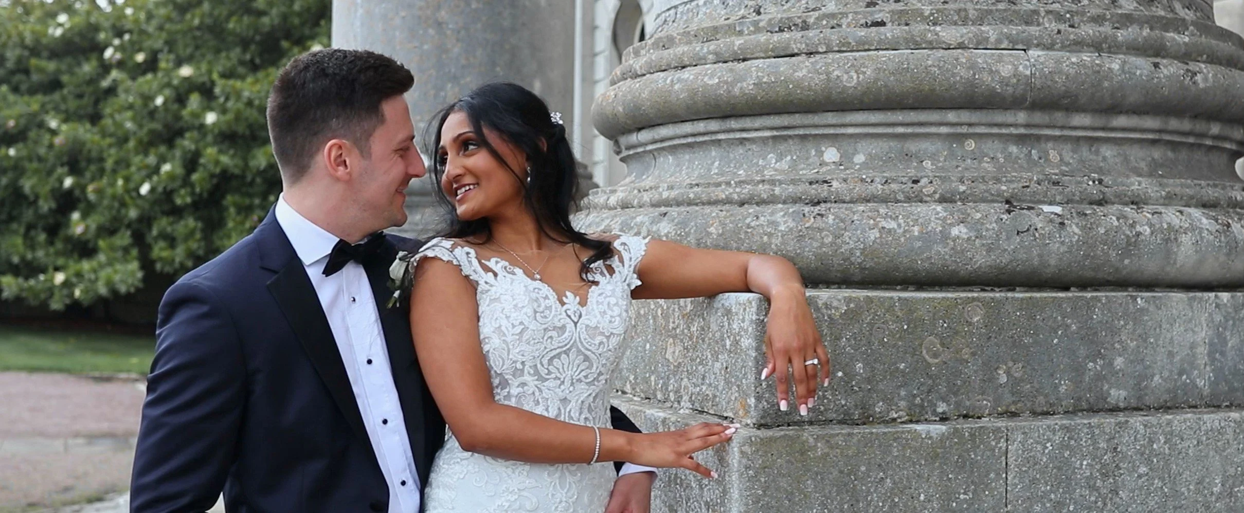 A newlywed couple smiling and gazing into each other's eyes next to a large stone monument, with greenery in the background.
