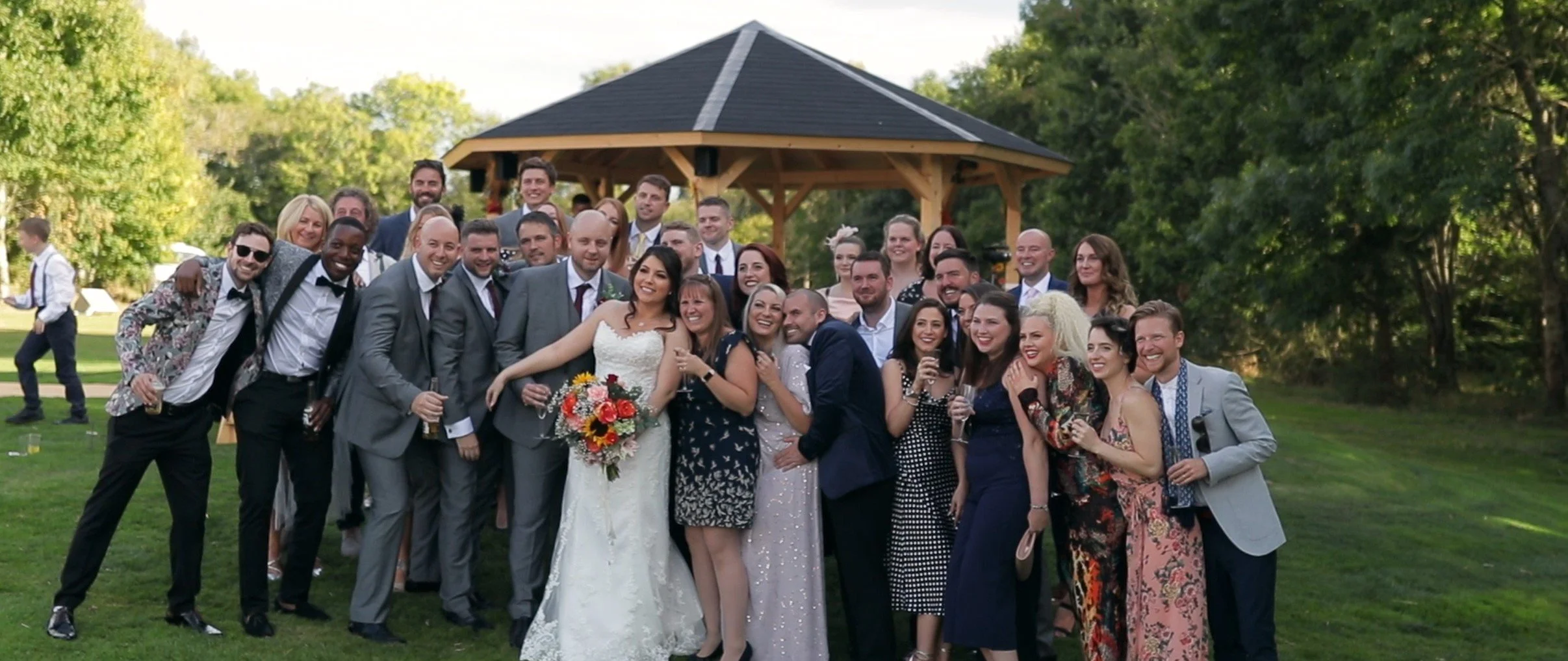 Group of wedding guests celebrating outdoors in the evening, smiling and socializing on a grassy area with trees and a gazebo in the background.