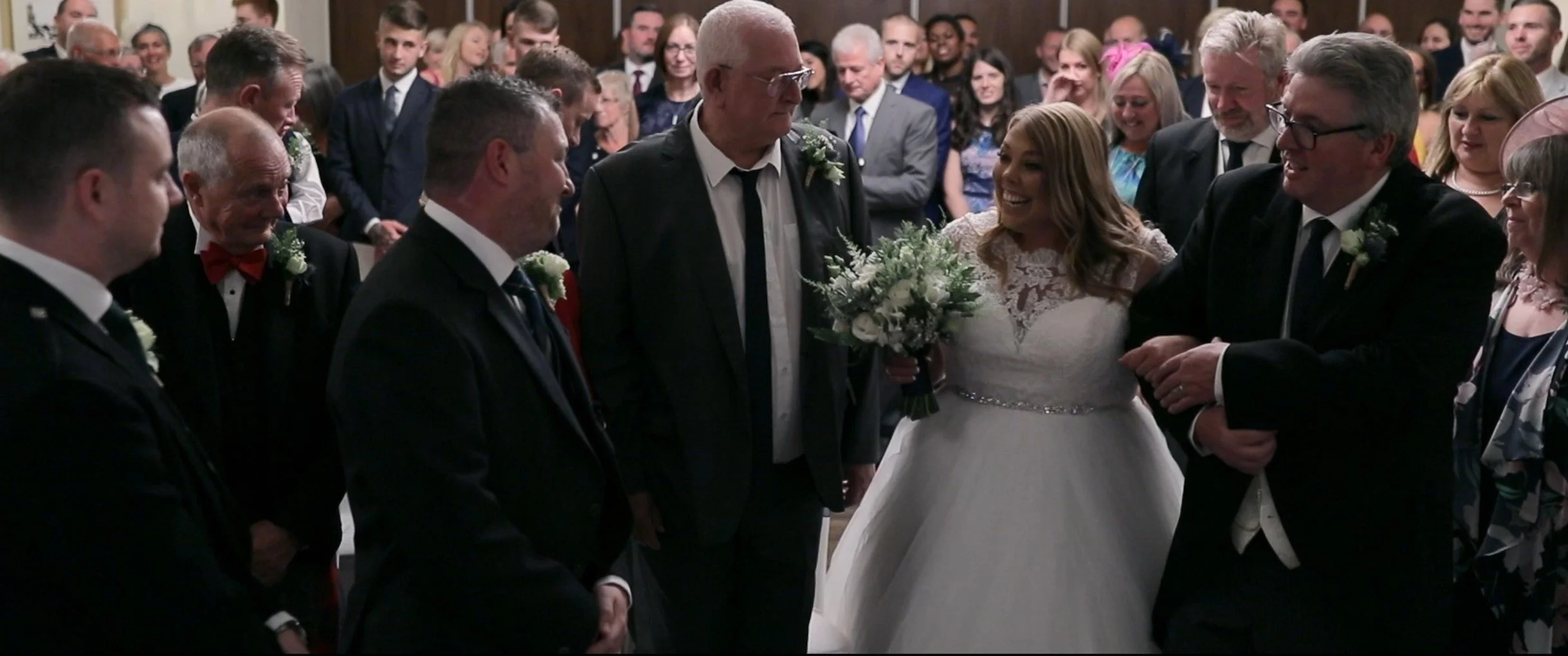 Bride in white wedding dress holding a bouquet of white flowers, standing among smiling guests at a wedding ceremony.