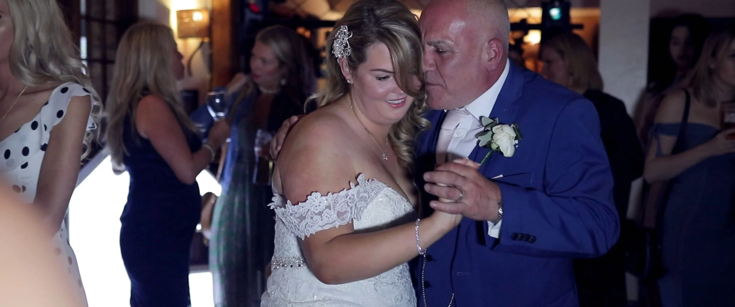 A bride and an older man, likely her father, dancing closely at a wedding reception. The bride is in a white lace off-the-shoulder wedding dress and the man is in a blue suit with a white shirt and boutonniere.
