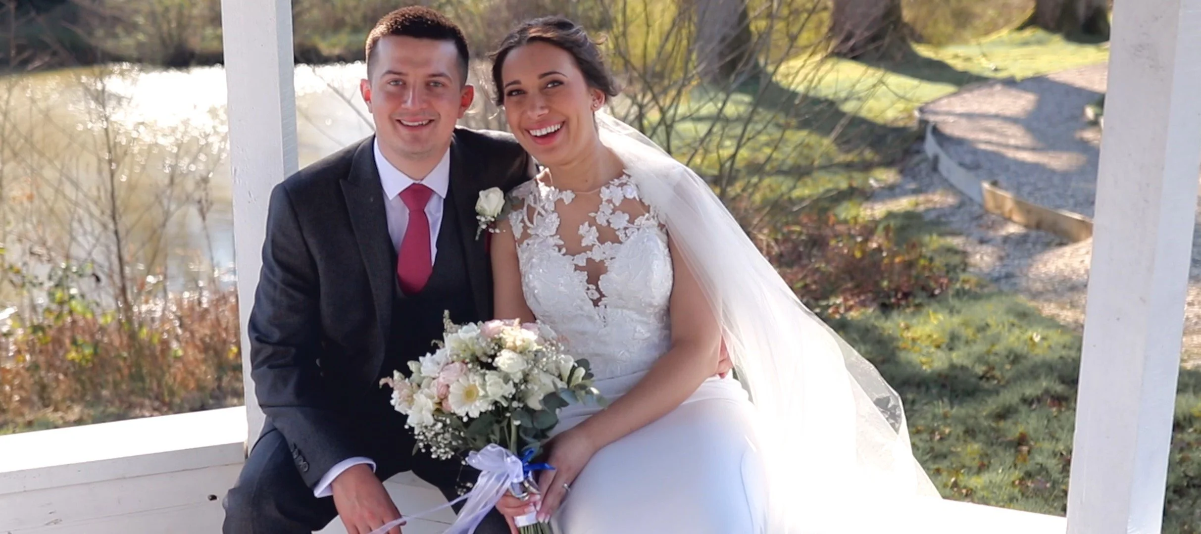 A newlywed couple sitting outdoors on a white bench, smiling at the camera. The groom is in a dark suit with a red tie, and the bride is in a white wedding dress holding a bouquet of white and pink flowers. The background features trees and a pathway