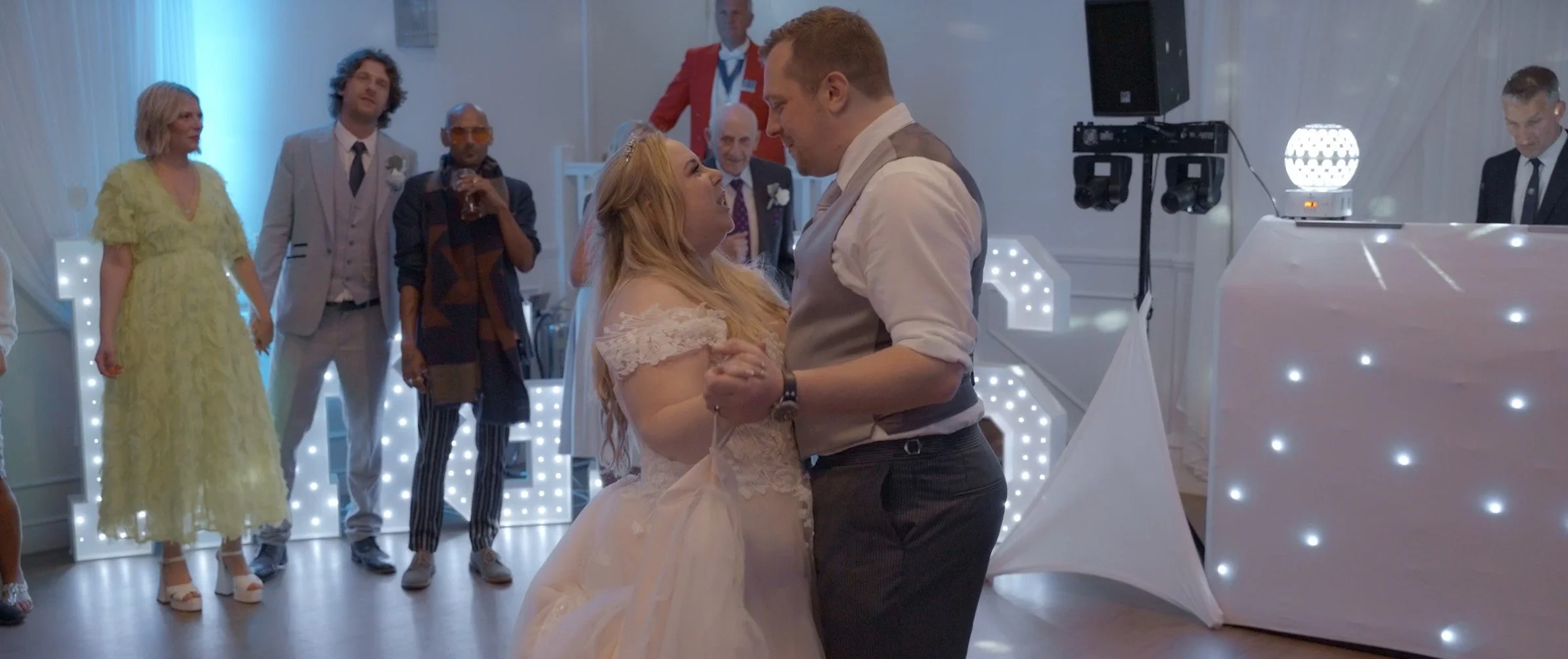 A wedding reception with a bride and groom dancing together as guests watch. The bride wears a light-colored wedding dress, and the groom wears a vest and shirt. Guests are dressed in formal attire, and the background features decorative lighting and