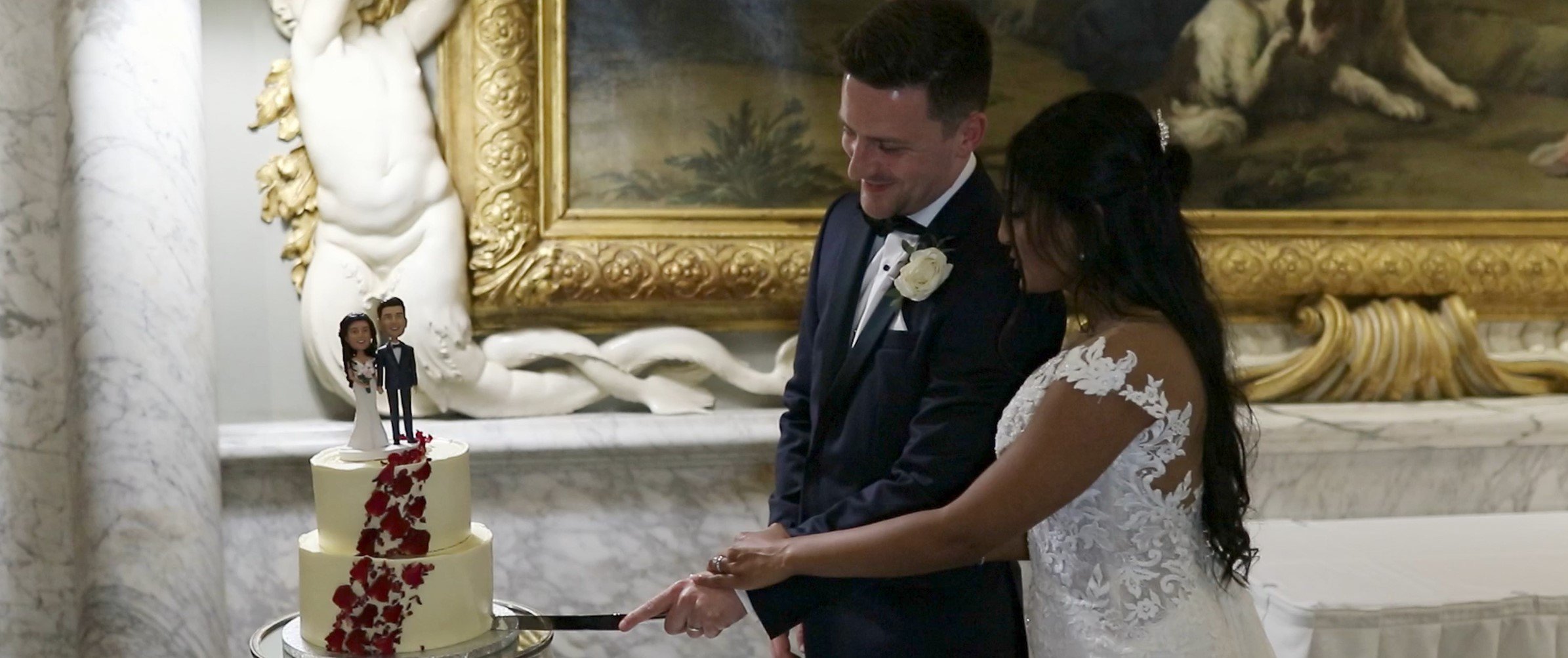 A bride and groom at their wedding cutting a two-tiered wedding cake with a topper of two cartoon-like figures dressed as a bride and groom, adorned with red flower decoration, in an ornate room with a large framed painting and decorative white marbl