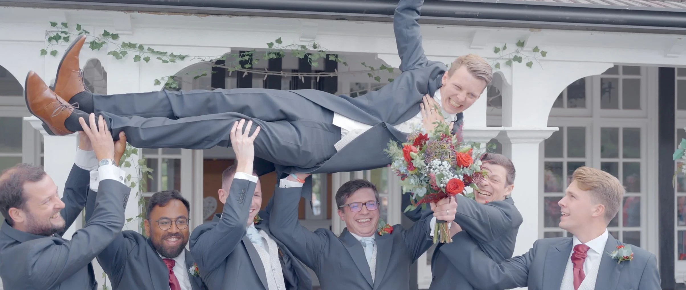A groom being lifted horizontally in the air by five men in suits at a wedding, holding a bouquet of flowers, porch of a house in the background.