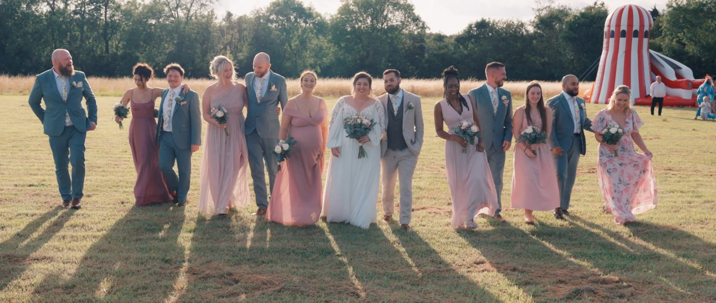 A diverse group of people in formal attire walking outdoors on a grass field during a sunny day, with a striped red and white inflatable slide in the background.