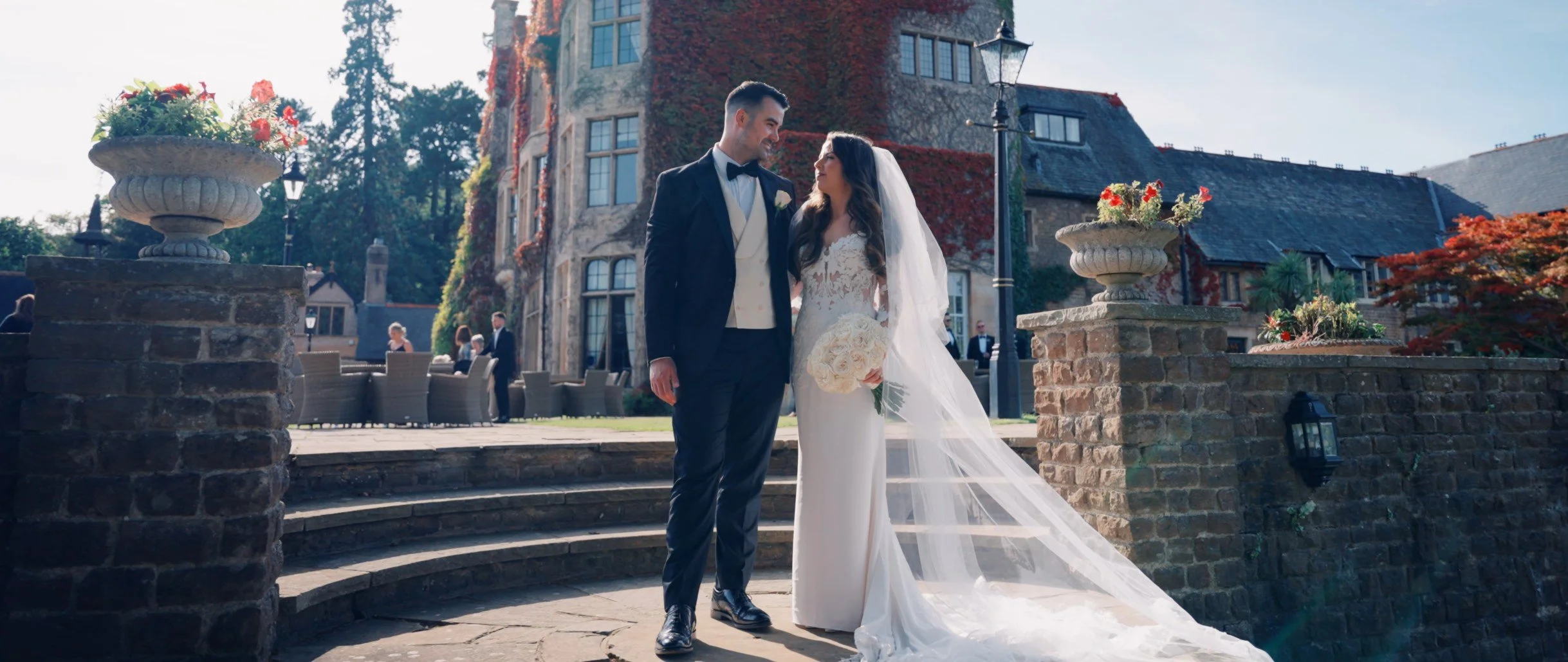 A bride and groom standing outside on a staircase in front of a historic building, sharing a moment on their wedding day. The bride wears a white gown with a long veil, holding a bouquet of flowers. The groom is in a dark tuxedo with a bow tie.