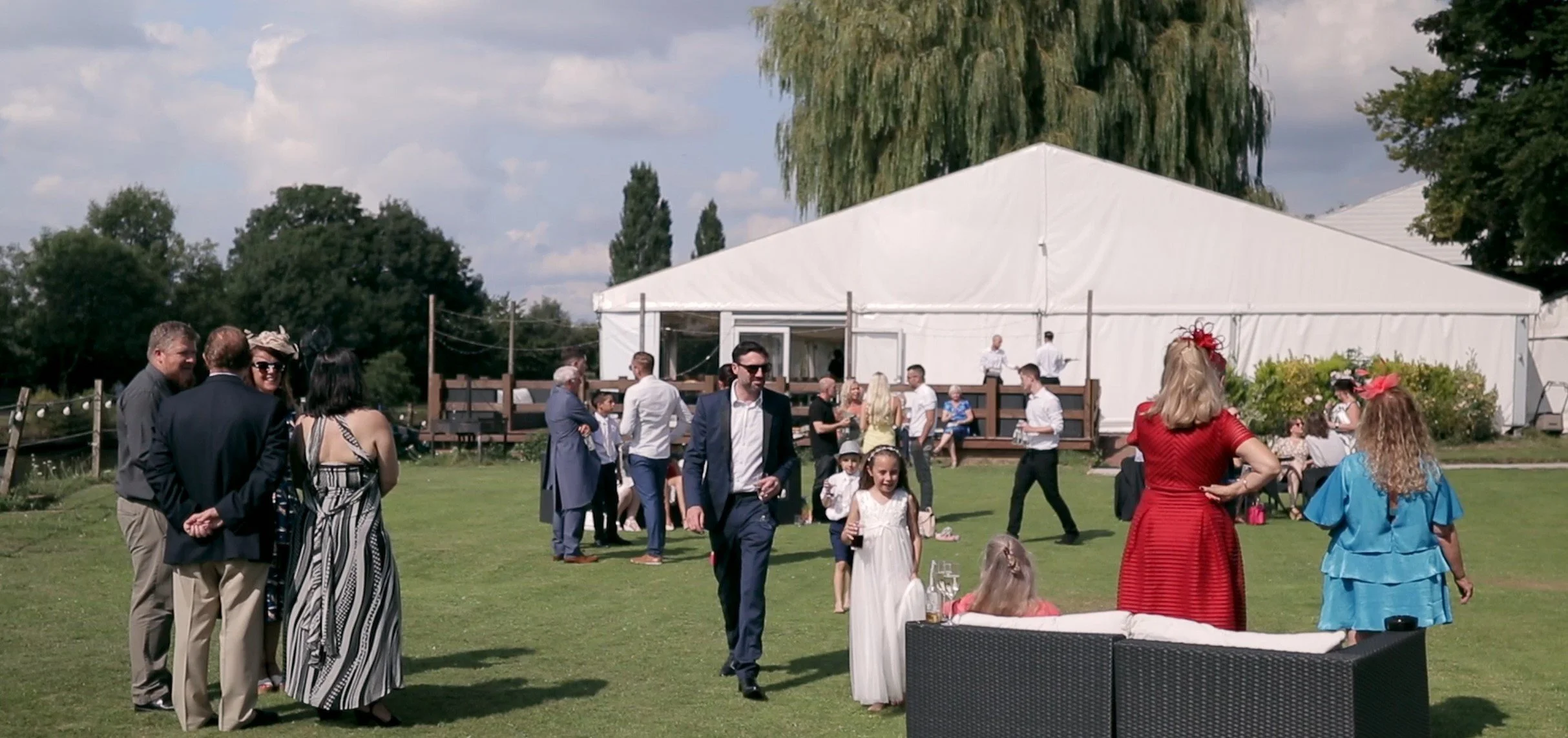 People socializing and children playing on a lawn at an outdoor event with a large white tent in the background under partly cloudy sky.