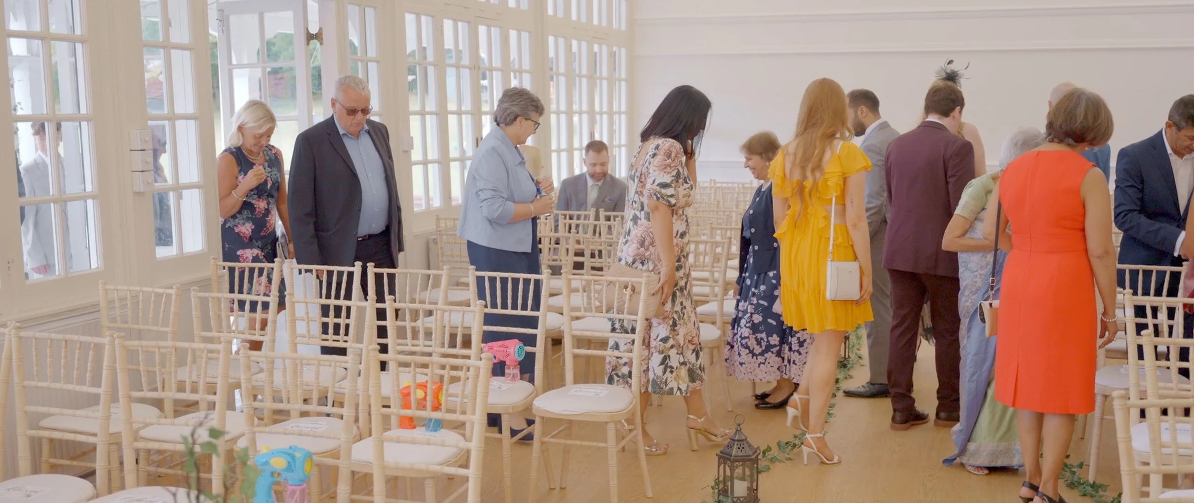 People gather inside a bright room with big windows, standing in a line or kneeling, dressed in formal and semi-formal attire, preparing for a ceremony.