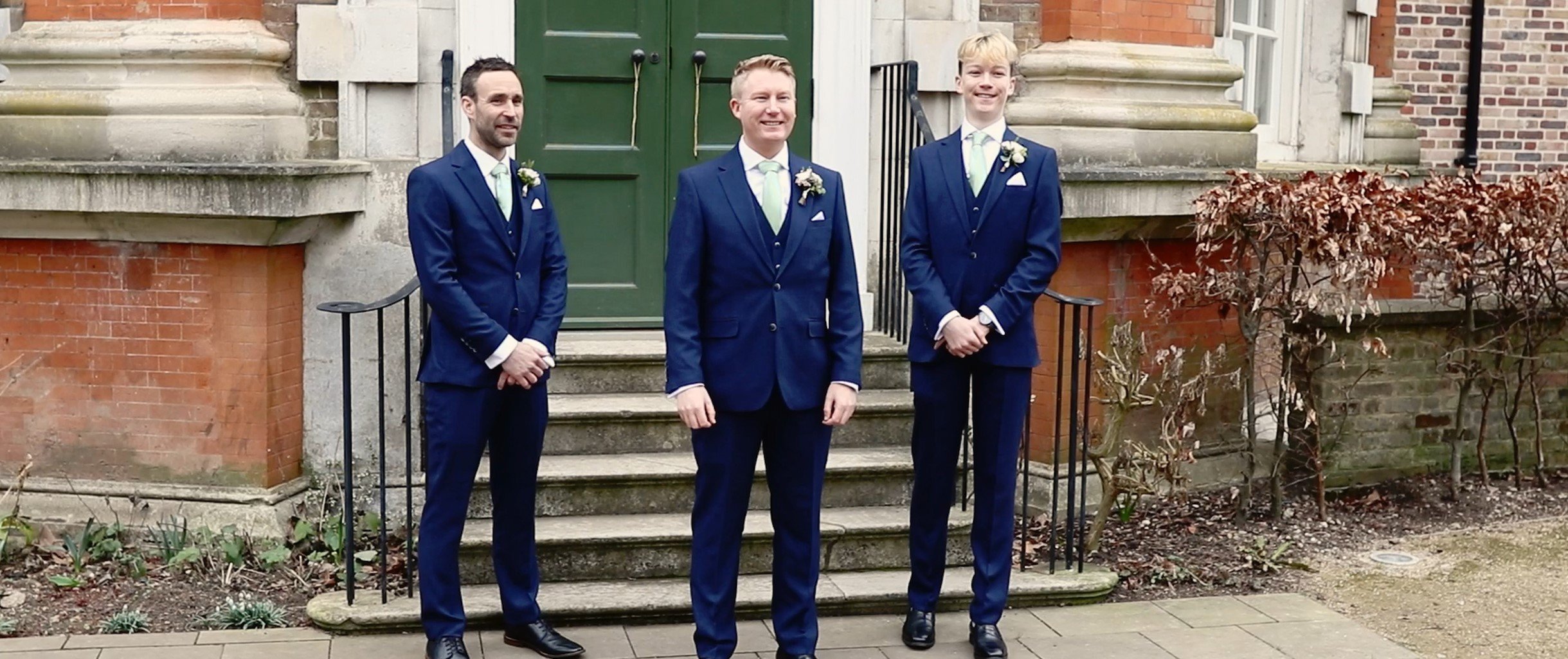 Three men dressed in navy suits with white shirts and green ties, standing on the steps outside a brick building with a green door. They are smiling and wearing boutonnières on their left lapels for a formal occasion.