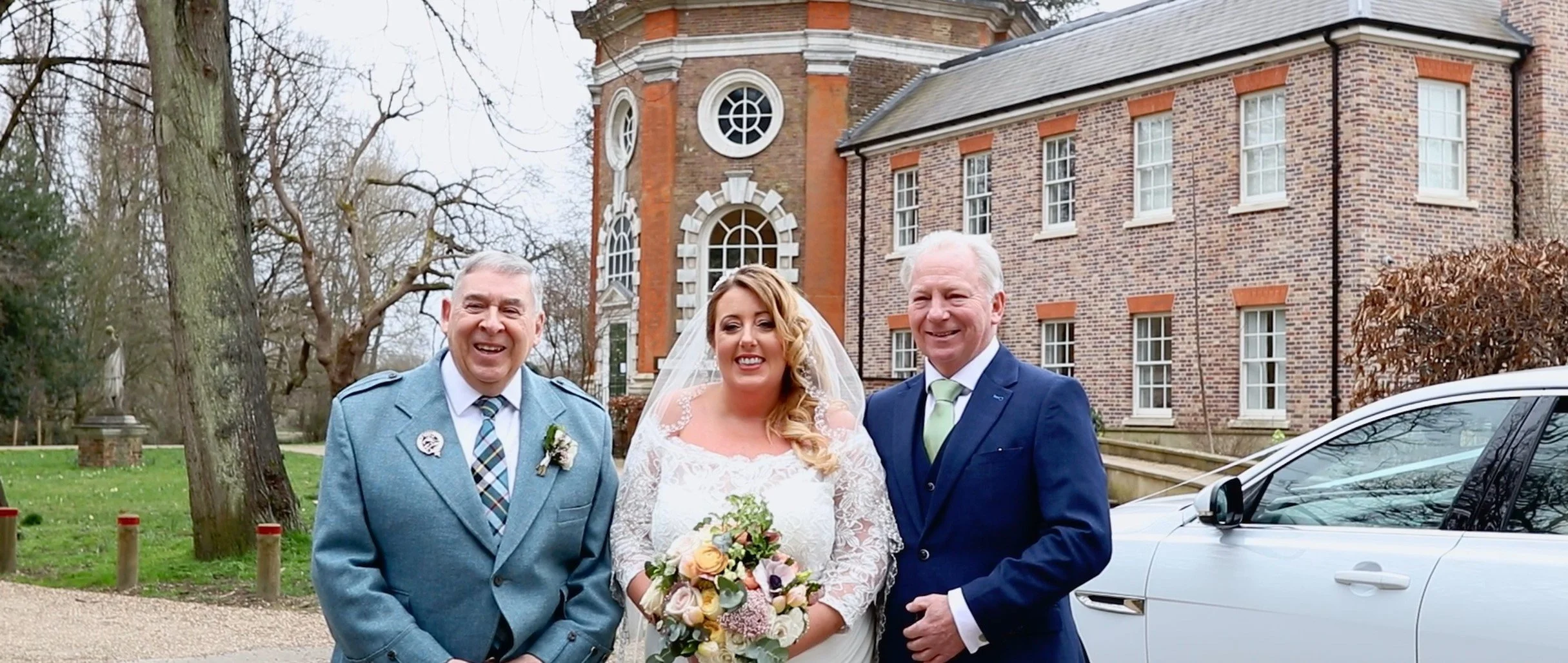 A bride in a white wedding dress holding a bouquet standing between two men in suits outside a brick building on a wedding day.
