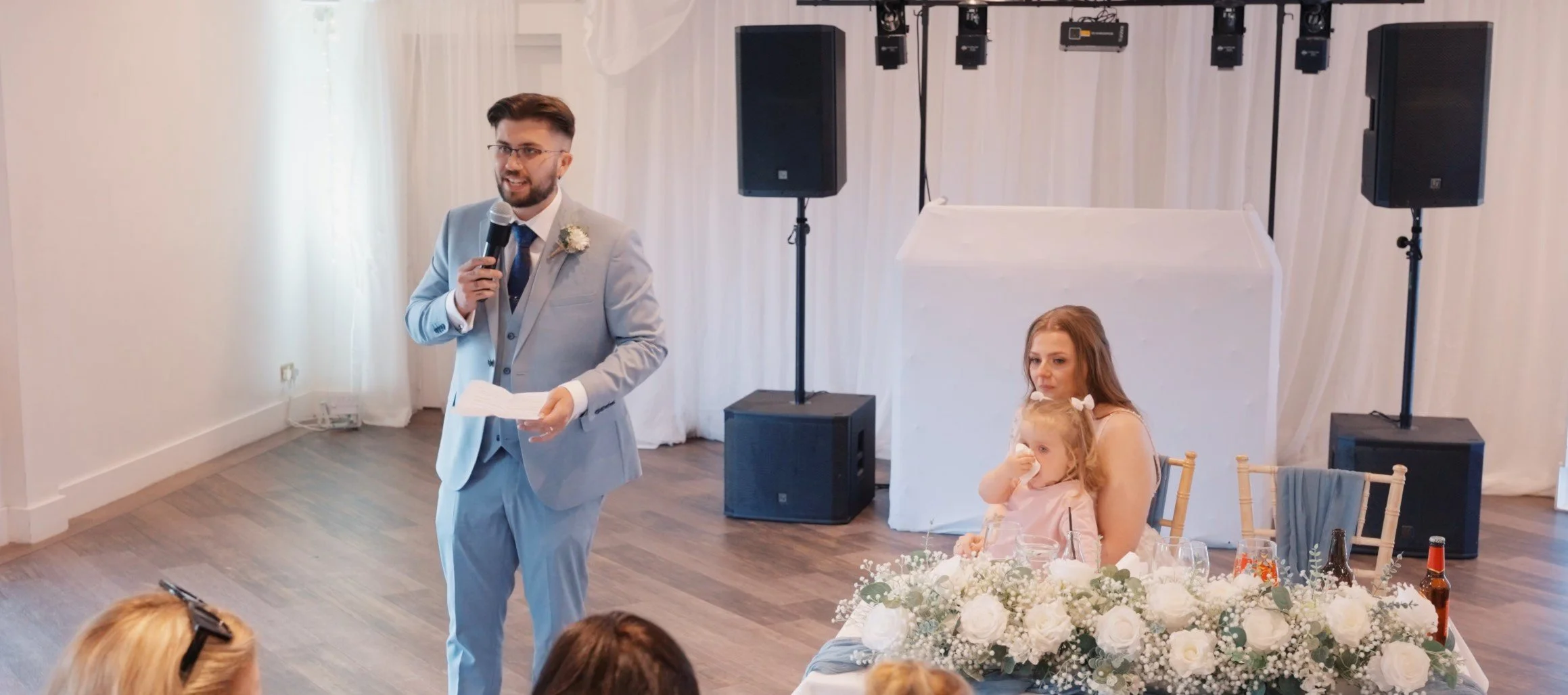 A man in a light gray suit with a blue tie and white shirt is giving a speech at a wedding reception, holding a microphone and some notes. There is a woman with red hair sitting next to a young girl with curly blonde hair, both sitting at a table dec