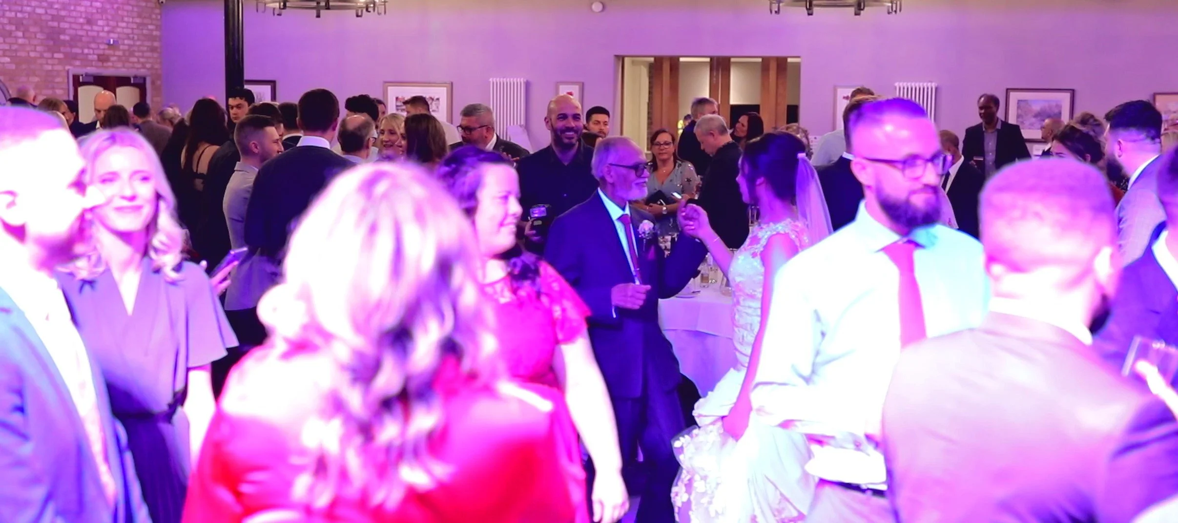 People at a social event or party, with some dancing and others chatting, in a decorated indoor venue with purple lighting.