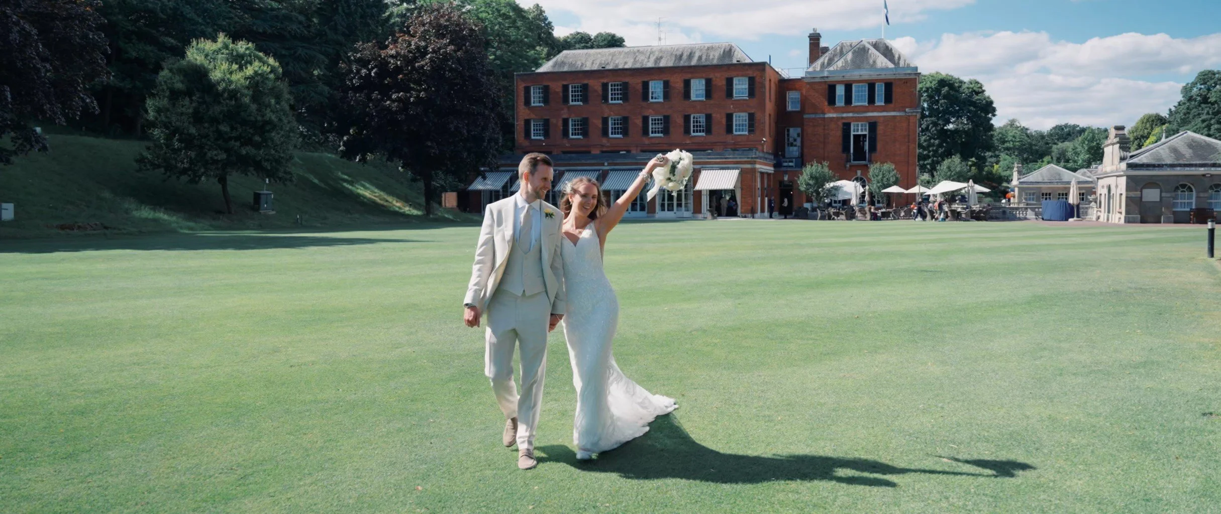 A bride and groom walking hand in hand on a green lawn during their wedding, with a large brick building and outdoor tables in the background on a sunny day.