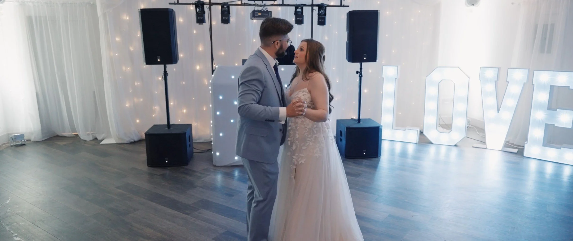 Couple dancing at wedding reception with large illuminated "LOVE" sign in background.