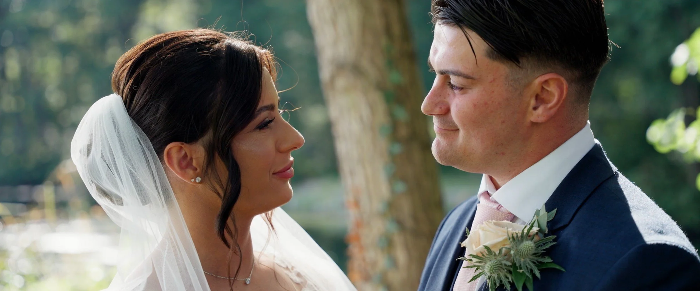 Bride and groom face each other outdoors on their wedding day, smiling softly with trees and sunlight in the background.