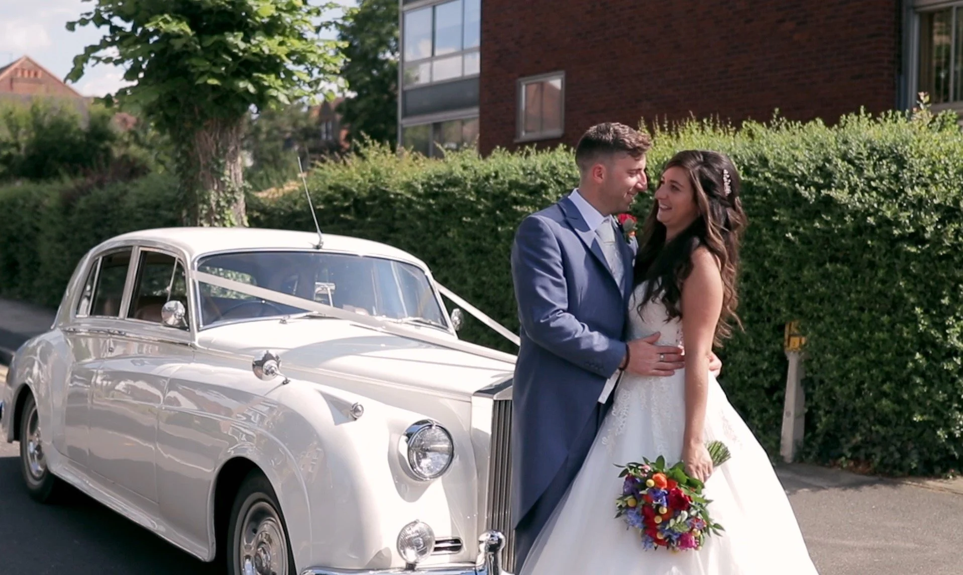 A newlywed couple in wedding attire in front of a vintage white car, smiling and holding each other, with a green hedge and brick building in the background.
