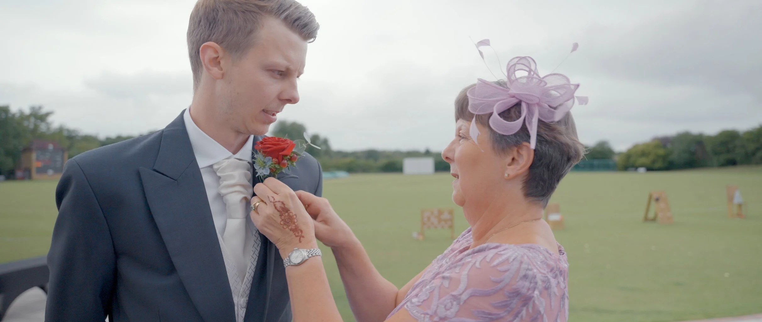 A woman in a pink dress and large pink hat pins a red boutonniere onto a young man's suit at an outdoor wedding.