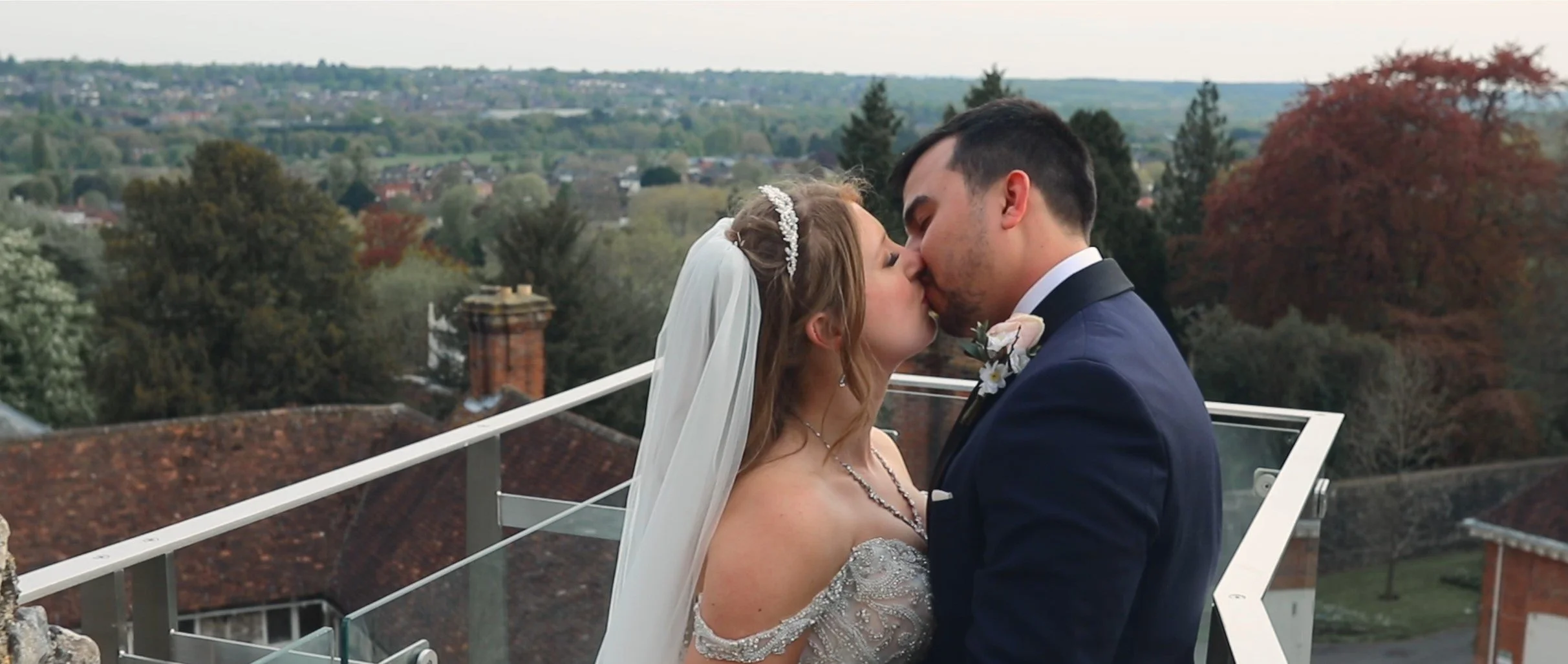 A bride and groom sharing a kiss on a rooftop, with trees and countryside in the background.