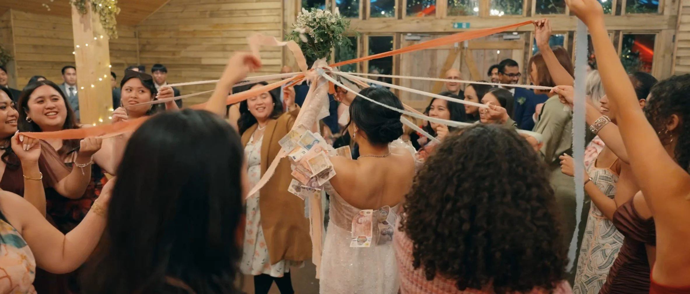 A bride in a wedding gown holding cash while surrounded by excited guests pulling ribbons attached to a central point, celebrating a wedding tradition at a festive event.
