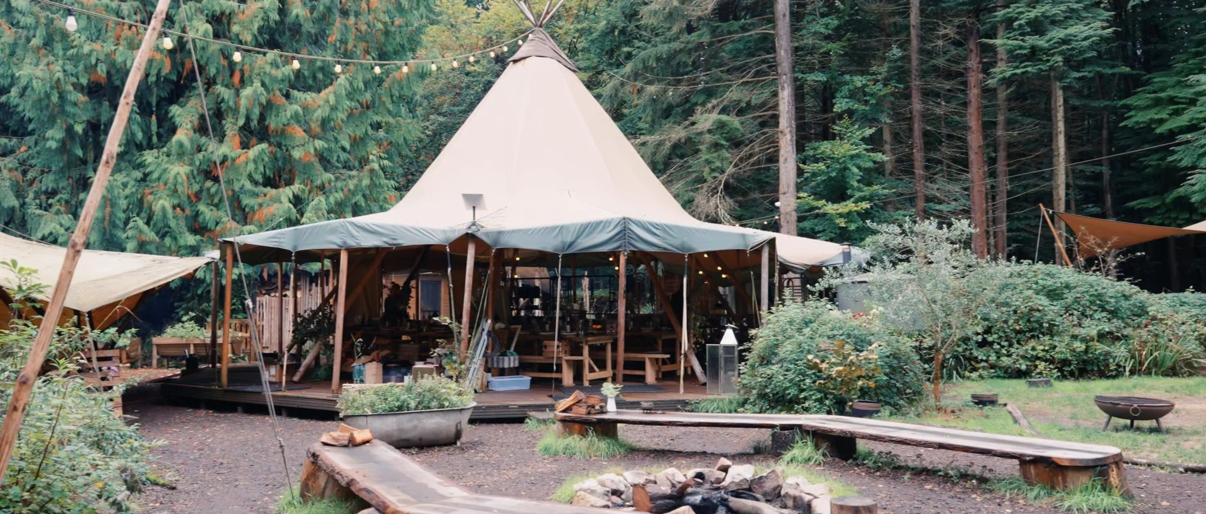 A large beige teepee tent set up in a forest, surrounded by green trees and bushes, with wooden benches and a fire pit in front.