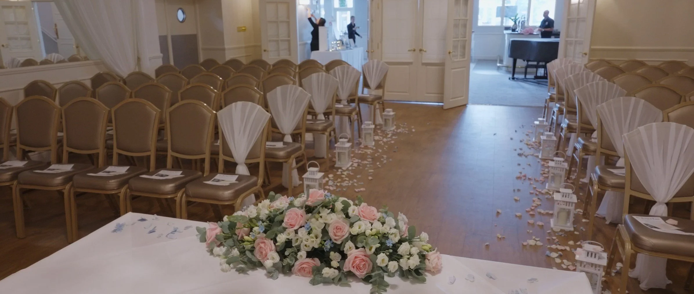 Empty wedding ceremony chairs decorated with white fans, white lanterns, and flower petals scattered on the floor, with a floral centerpiece in the foreground, inside a decorated room.
