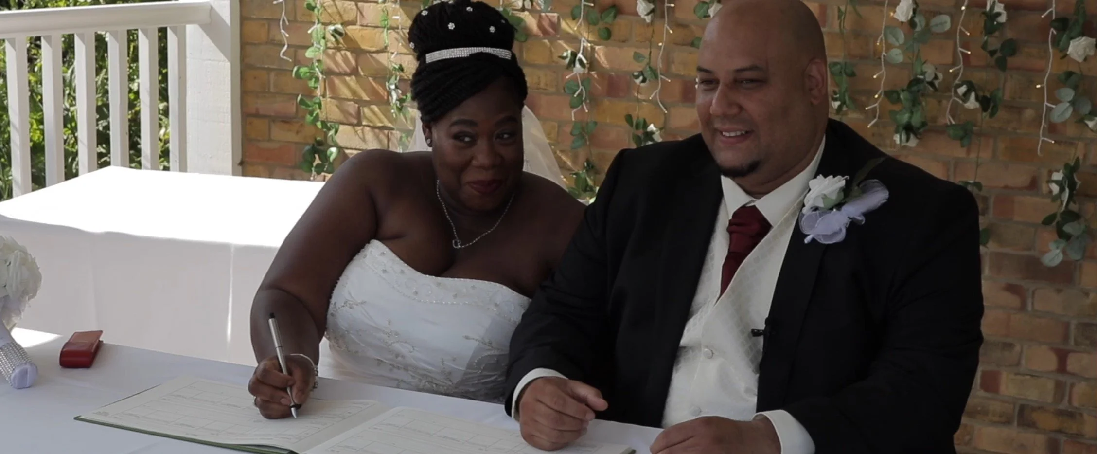 A bride and groom sitting at a table during their wedding ceremony. The bride, wearing a white dress, is signing a document, while the groom, in a black suit with a white shirt and red tie, smiles.