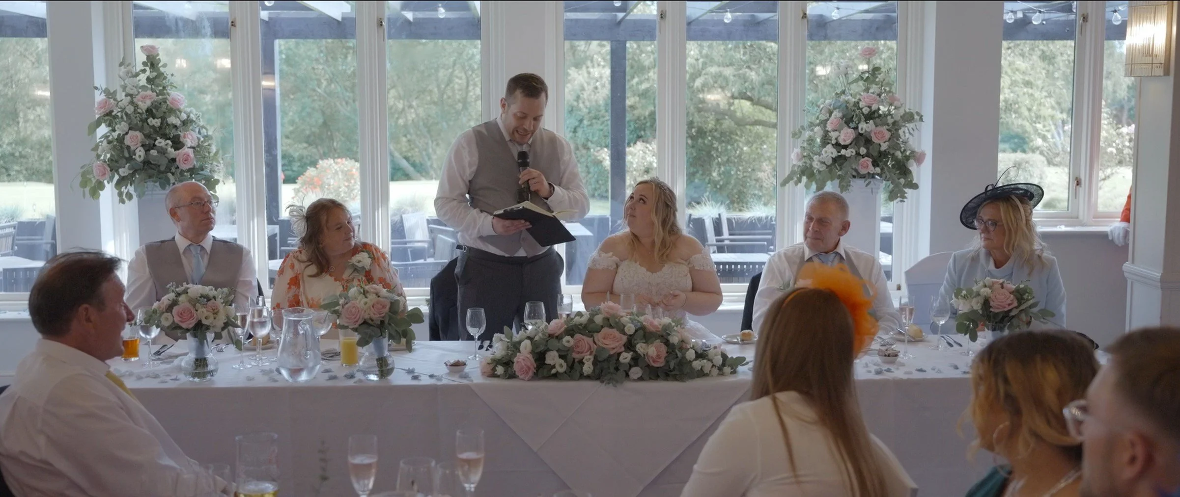 A man giving a speech at a wedding reception table with four women and two men, decorated with pink and white flowers, inside a bright room with large windows and outdoor view.