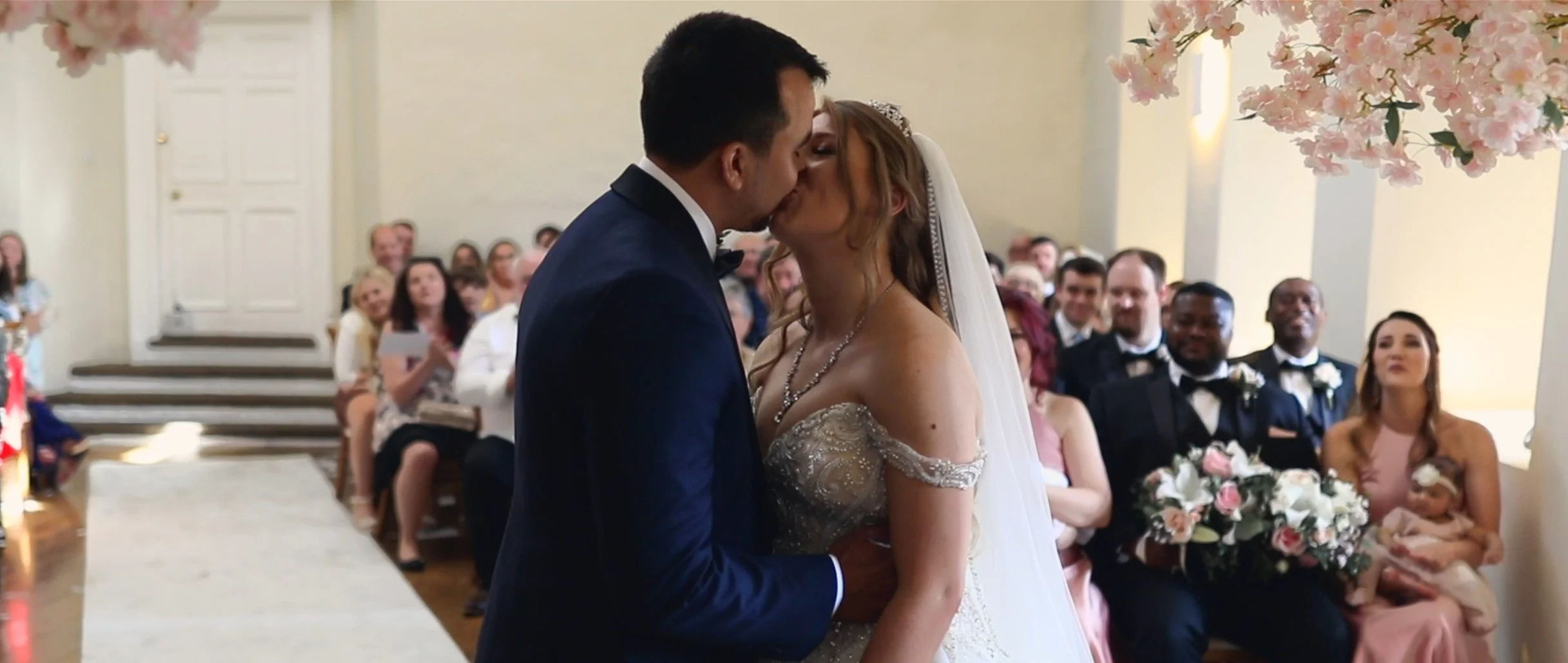 A bride and groom share a kiss during their wedding ceremony with wedding guests seated in the background, decorated with pink floral arrangements.