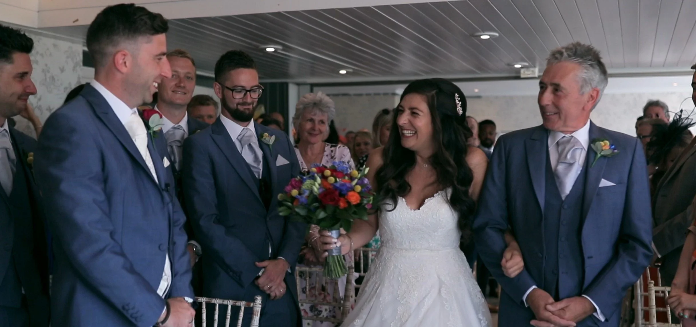 Bride in a white wedding dress holding a bouquet of colorful flowers, standing amidst groomsmen in blue suits, smiling and looking at her during a wedding ceremony.
