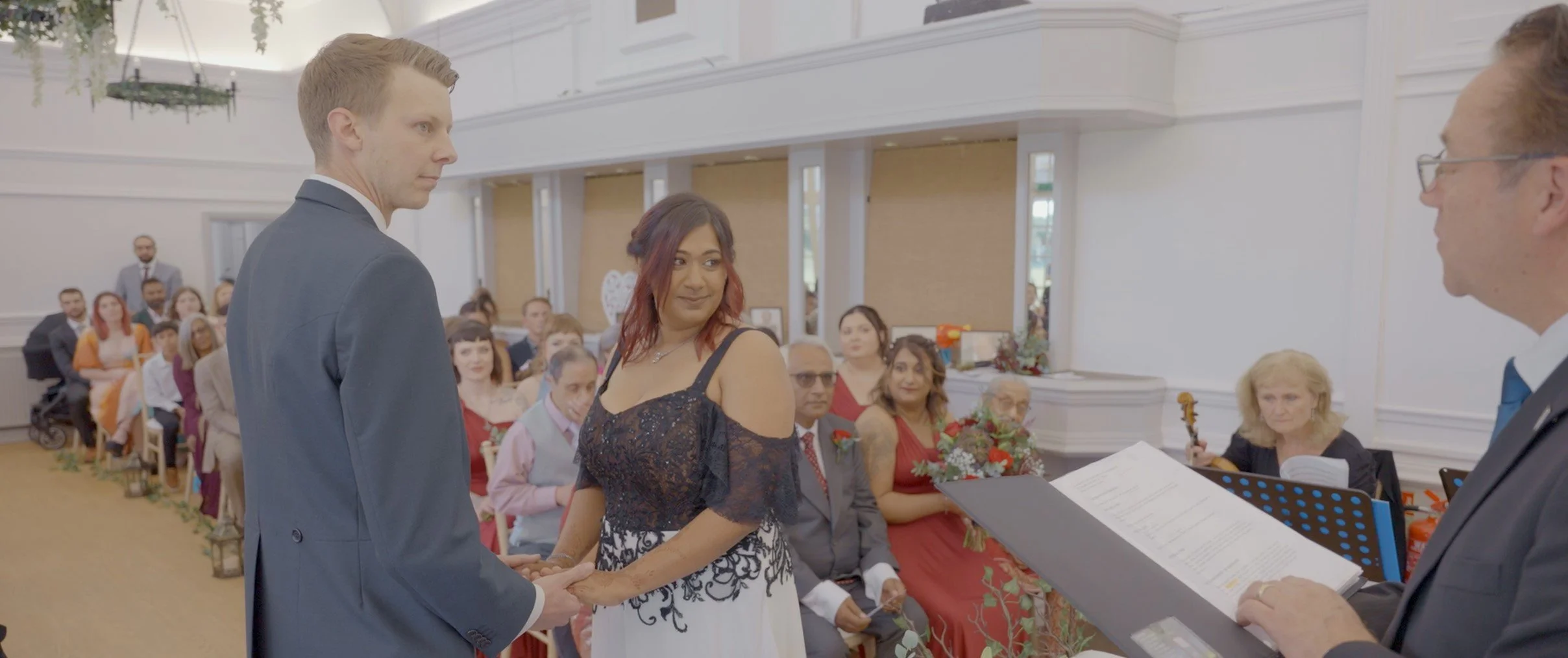 A wedding ceremony where a man and woman are exchanging vows. The woman has reddish-brown hair and is wearing a black and white dress, while the man has light hair and is dressed in a suit. They are facing each other, holding hands, with guests seate
