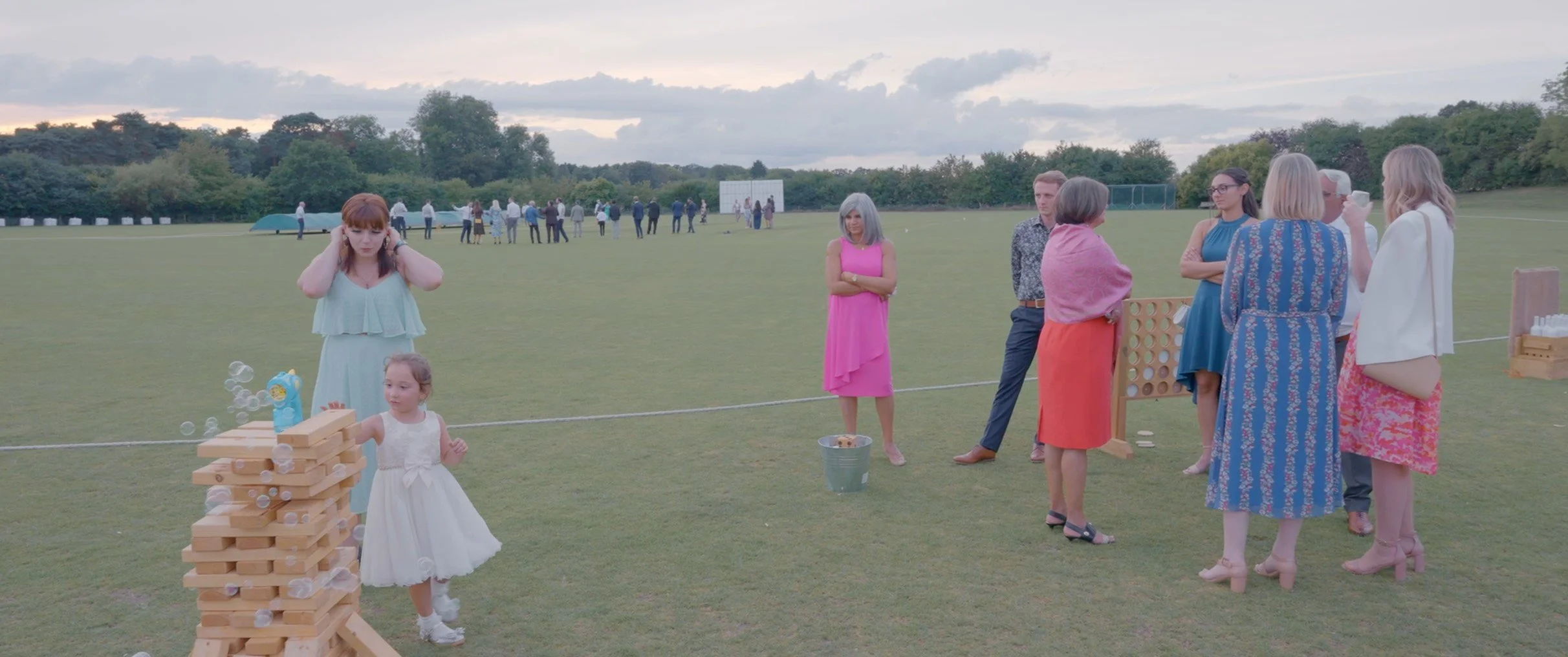 People gathered outdoors on a grassy field during daytime, with women and children playing a game of giant Jenga, and other guests socializing.