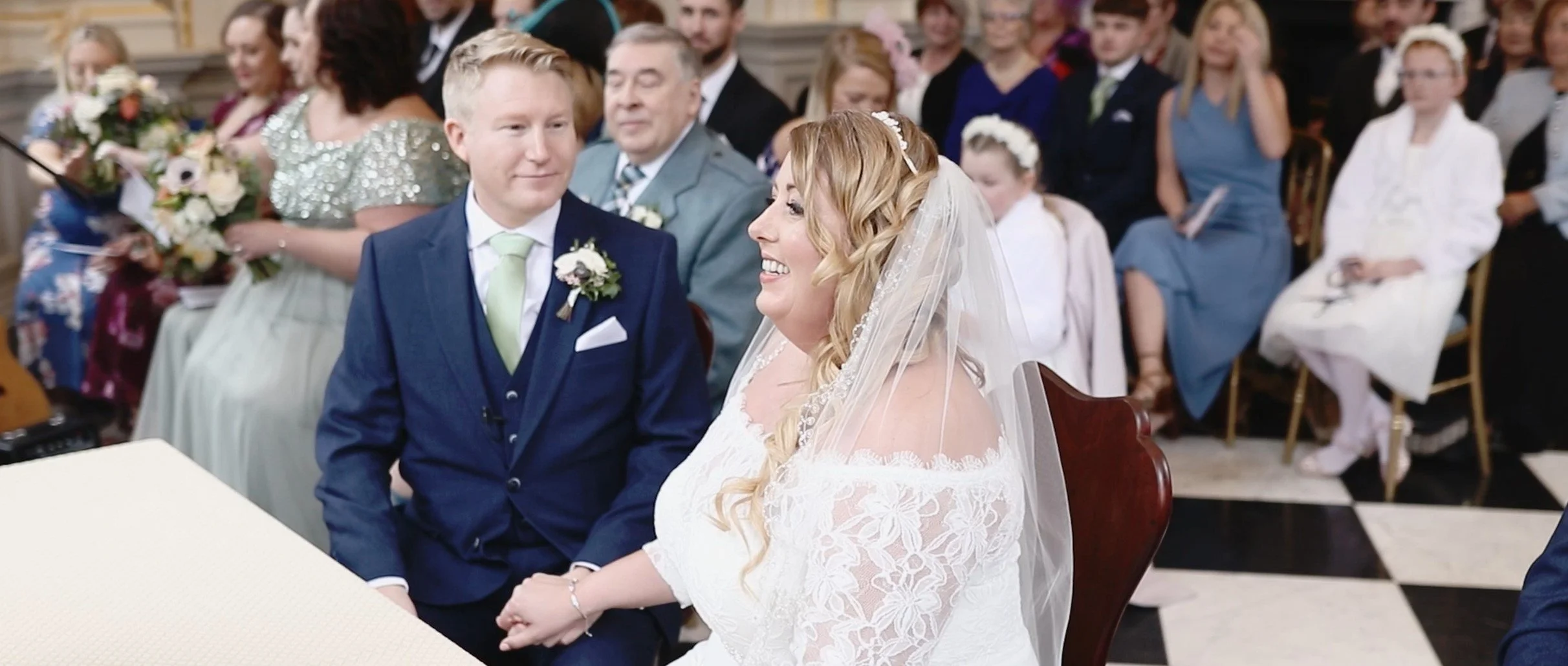 Bride and groom during wedding ceremony, holding hands, with guests seated behind them in a church or wedding venue.