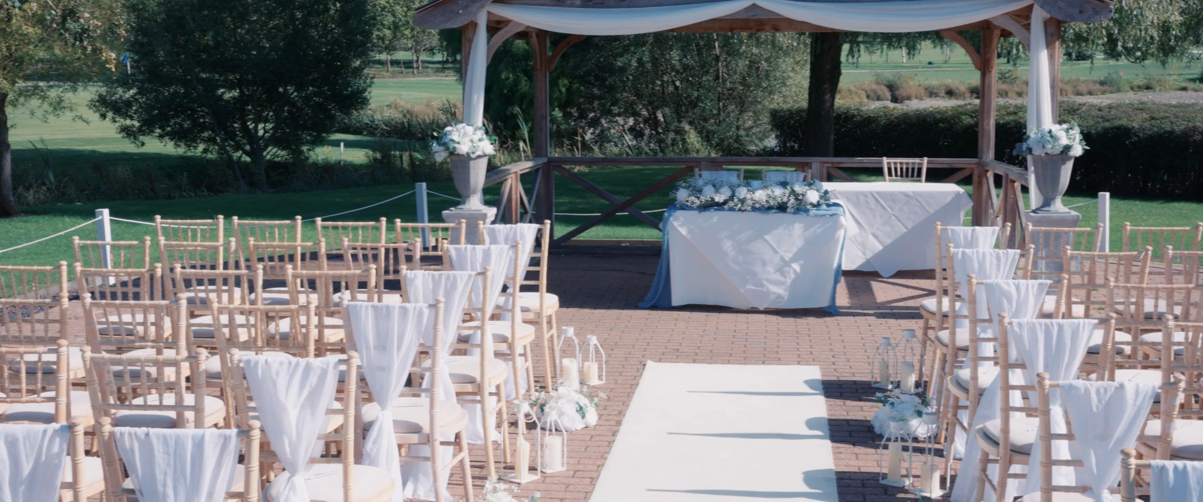 Outdoor wedding ceremony setup with rows of chairs facing a decorated altar under a gazebo, with floral arrangements and lanterns on a brick-paved area.
